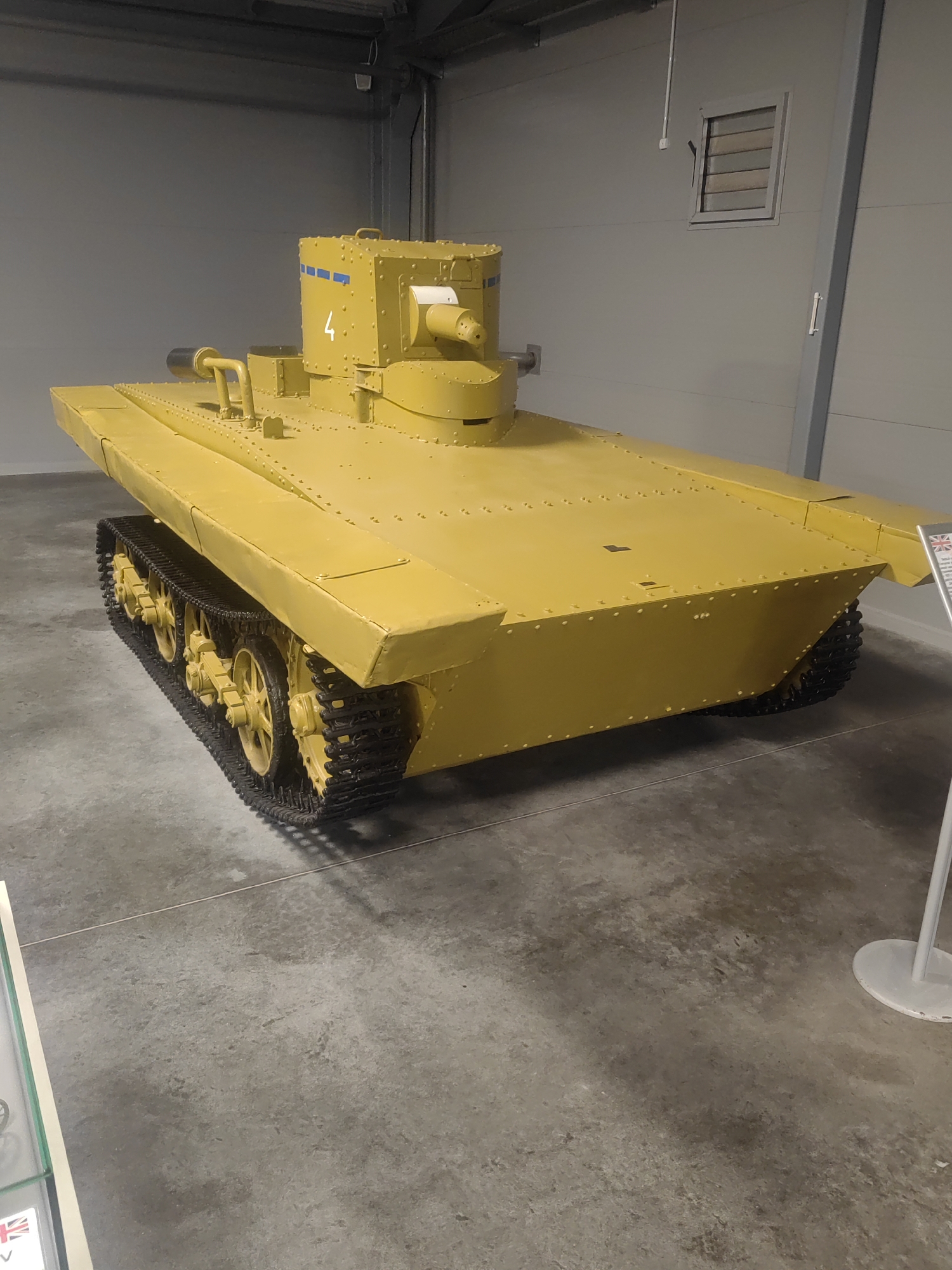 This is a media file of a yellow tank displayed indoors. The tank is the main subject of the image, and it appears to be a historical artifact. The setting is a museum or exhibition hall, with concrete flooring and plain, grey walls. There is a white vent on the wall in the background, and some lighting fixtures are visible on the ceiling. A white pole with a round base is present to the right of the tank.
The tank itself is bright yellow and has a distinctive design with a long, flat hull and a small turret on top. The turret has a gun protruding from the front and is marked with the number "4" in white and blue stripes. The tracks of the tank are black. There are many rivets visible on the hull and turret, suggesting it is an older model.
To the far right of the image, a small portion of a display sign is visible, featuring the Union Jack flag and some text, suggesting the tank may be British. The overall atmosphere is neutral, typical of a museum display. There is no indication of the time of day or weather conditions as the scene is indoors. No people are visible in the image.
