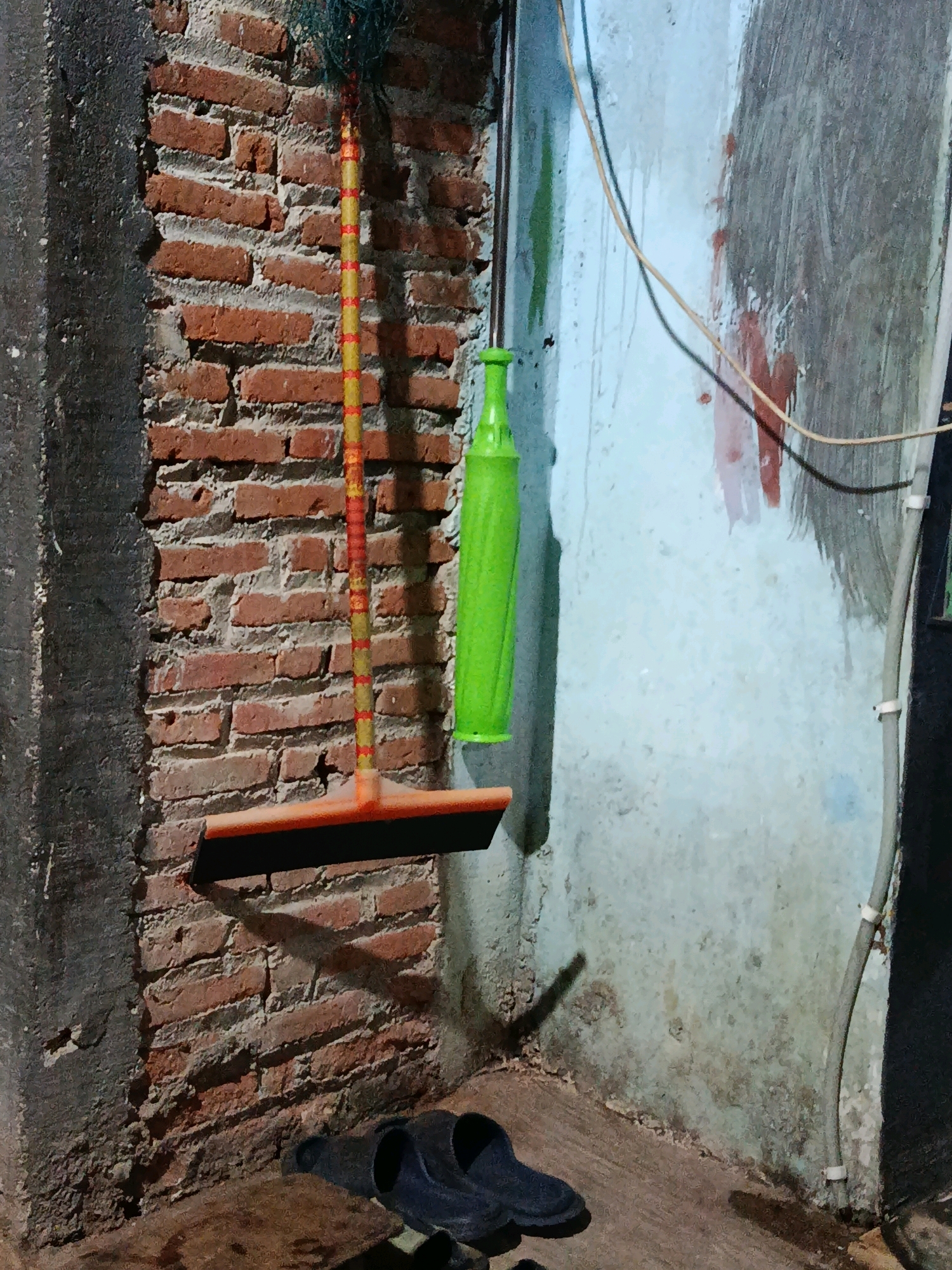 In a dimly lit corner of a building, possibly a storage area or a humble residence, in Jepara, Indonesia, a floor broom with an orange rubber squeegee head and a red and yellow striped handle hangs vertically against a rough brick wall. To its right, a bright green bottle, possibly a cleaning product or a water bottle, is also suspended. The wall is weathered and stained, with peeling paint and water marks, suggesting it may be an exterior wall or a damp interior space. Electrical wires run vertically along the wall, secured by white clips. In the foreground, a pair of dark, simple slippers rests on the floor, which appears to be a concrete or dirt surface. The scene conveys a sense of utilitarian simplicity, common in many Indonesian homes or workshops.