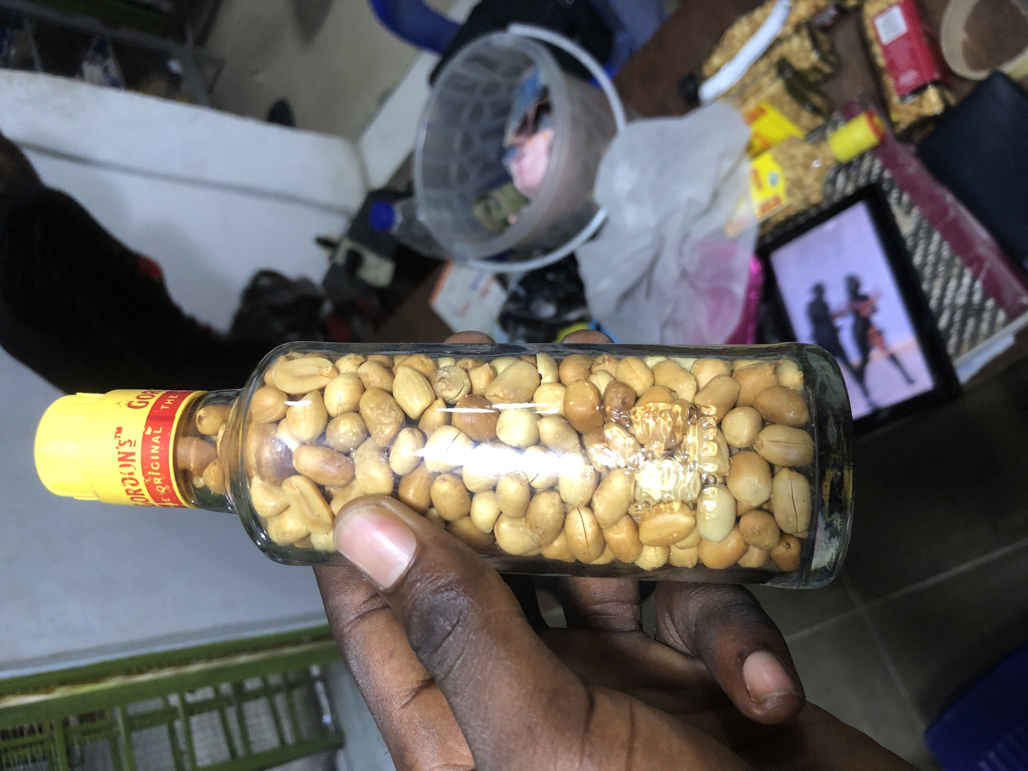 A hand, exhibiting dark skin and visible fingernails, holds a clear glass bottle filled with peanuts. The bottle has a yellow cap with red text that reads "GORDON'S" and "THE ORIGINAL". The background is a dimly lit interior, possibly a room or a storage area. Objects visible include shelves, a blue chair, a clear plastic bucket containing various items, and what appears to be a tablet device displaying an image of two people. The overall environment suggests an indoor setting in Ado, Nigeria, with the presence of consumer goods and everyday items.