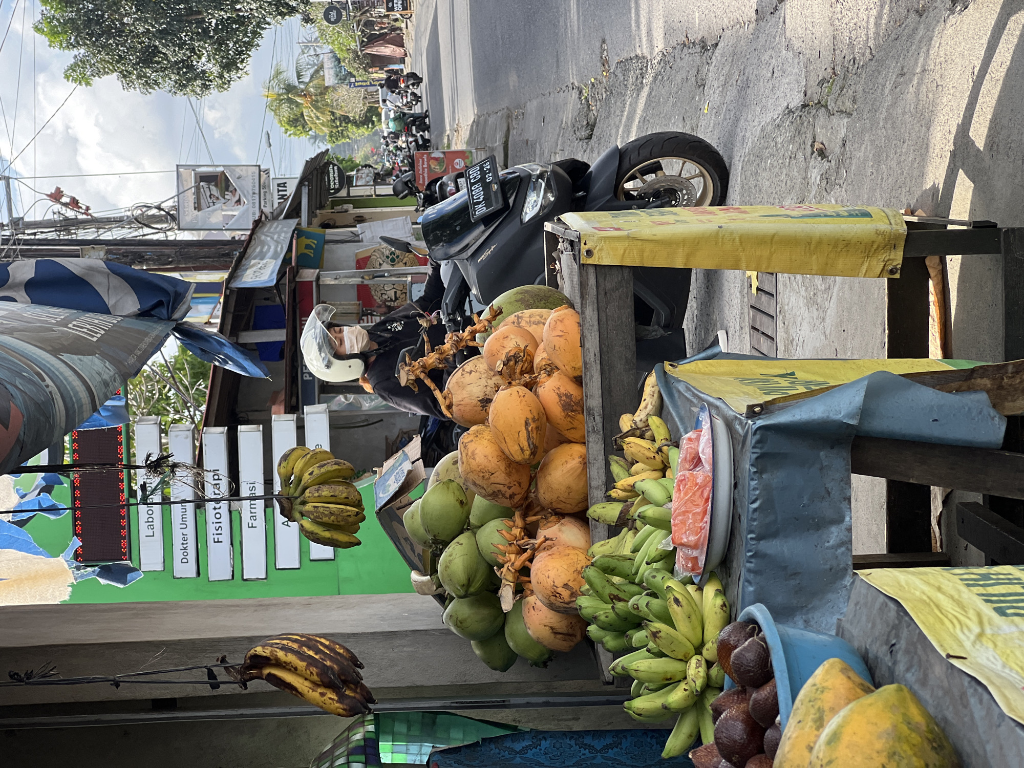 The image shows a roadside fruit stand in what appears to be Ubud District, Indonesia. The stand displays coconuts, bananas of varying ripeness, papaya, and salak fruit. A motorcycle is parked next to the stand, its license plate visible. Across the street, various business signs are visible, including a sign for "Laborator," "Dokter Umum," "Fisioterapi," and "Farmasi" above "Parking 50." A sign indicating a food place such as "Smoothie Bowls" can be seen near parked motorbikes.
