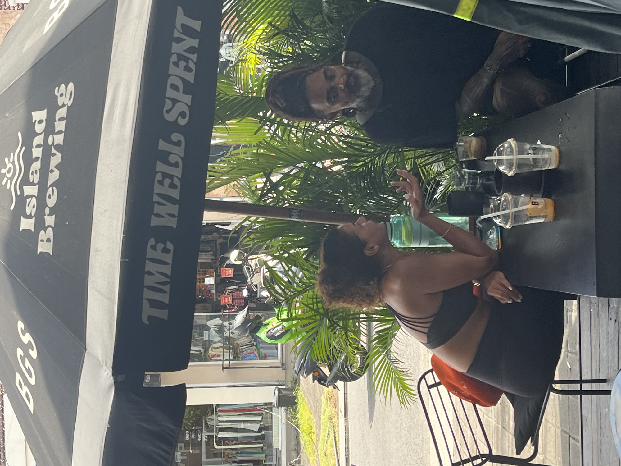 Two people, a man and a woman, are sitting at a black table under a large black umbrella that reads "BGS Island Brewing TIME WELL SPENT." The woman, with curly brown hair and wearing a black sports bra and black leggings, is seated on the left, gesturing with her right hand. She has a bright orange bag on the chair next to her. In front of her are two clear plastic cups with straws, one with an orange drink and the letter "B" visible, and the other with some residue. A green bottle is also on the table. The man, with dreadlocks and a beard, is seated on the right, wearing a black long-sleeved shirt. He has tattoos on his arms and is looking towards the woman. Behind them, lush green palm fronds create a tropical backdrop. In the background, a street scene is visible, with shops displaying clothing on racks and a person on a green motorcycle wearing a helmet and a bright green jacket. The environment appears to be an outdoor seating area of a cafe or bar in North Kuta, Indonesia, suggested by the "Island Brewing" text on the umbrella and the general tropical aesthetic.