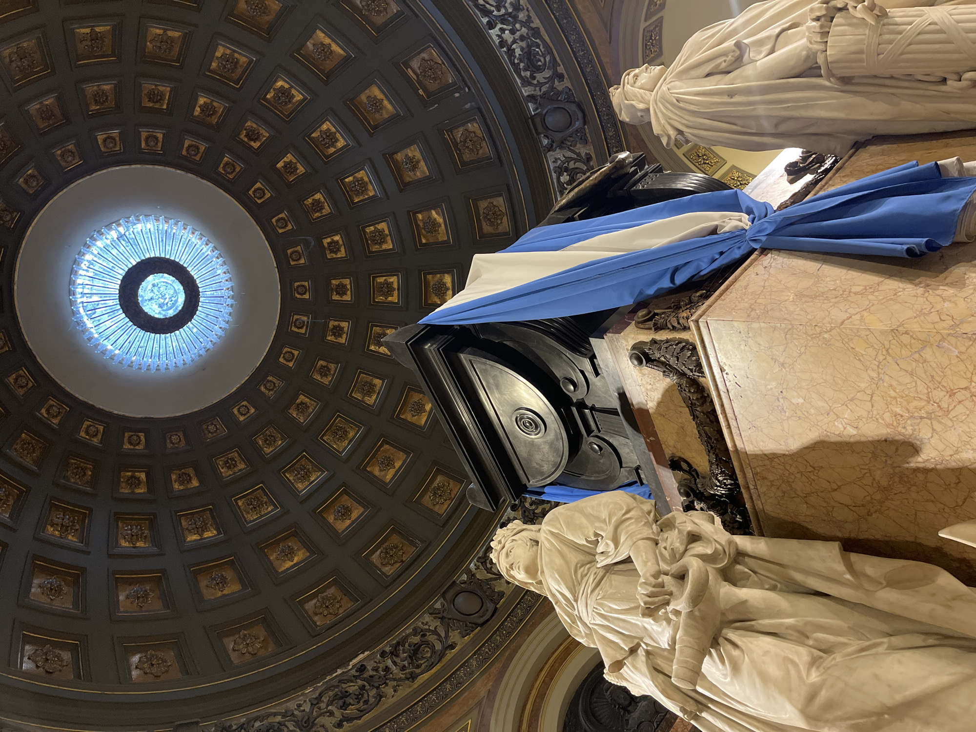 The image depicts a view upward inside a building in Buenos Aires, Argentina. A marble statue of a person lying down rests on a marble structure. The Argentinian flag is draped around it. The ceiling is a gold and black dome with a central light fixture.