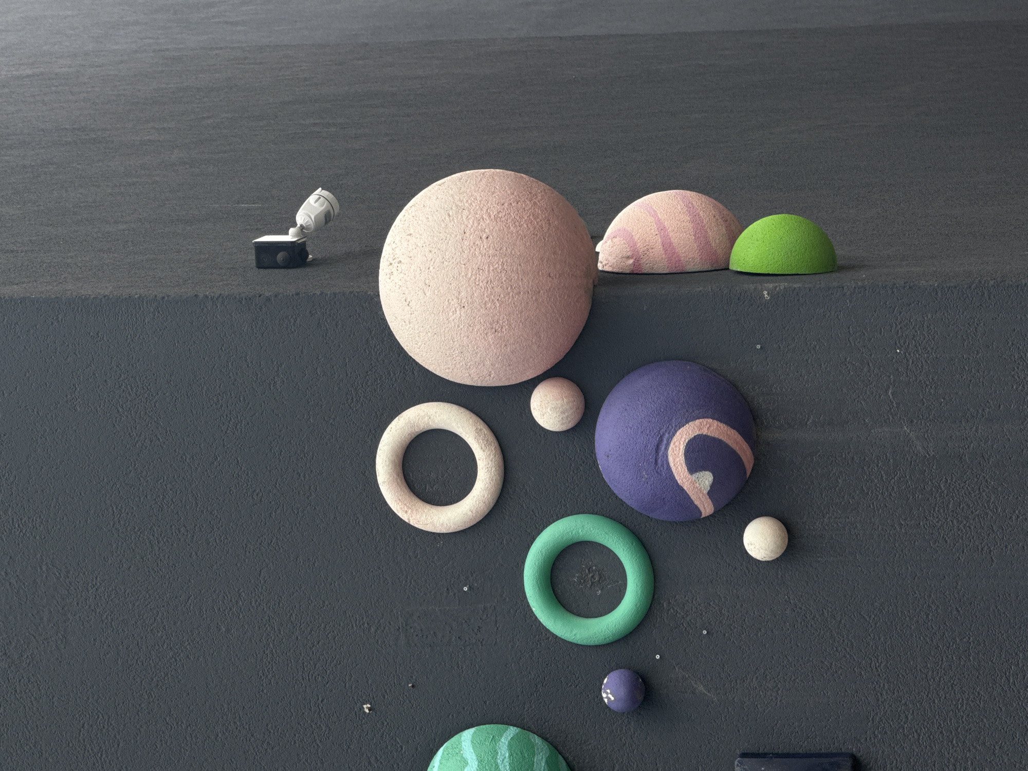 The image depicts a minimalist, abstract arrangement of various spherical and ring-shaped objects on a textured, dark gray surface. On a raised platform in the upper left, a small, white spotlight is aimed downwards. To its right, on the same platform, sit a large, light pink sphere with a textured surface, a light pink and white striped dome, and a bright green dome. Below the platform, on the dark gray surface, several more objects are arranged: a light-colored ring, a vibrant purple sphere with a light pink swirl design, a small white sphere, a teal-colored ring, a small purple sphere, and a portion of a green and white striped sphere at the bottom edge of the frame. There is no indication of people, weather, time of day, or emotions. The setting is abstract and lacks specific location cues beyond the provided context of Yekaterinburg, Russia, which is not visually represented in the image. No visible text is present.