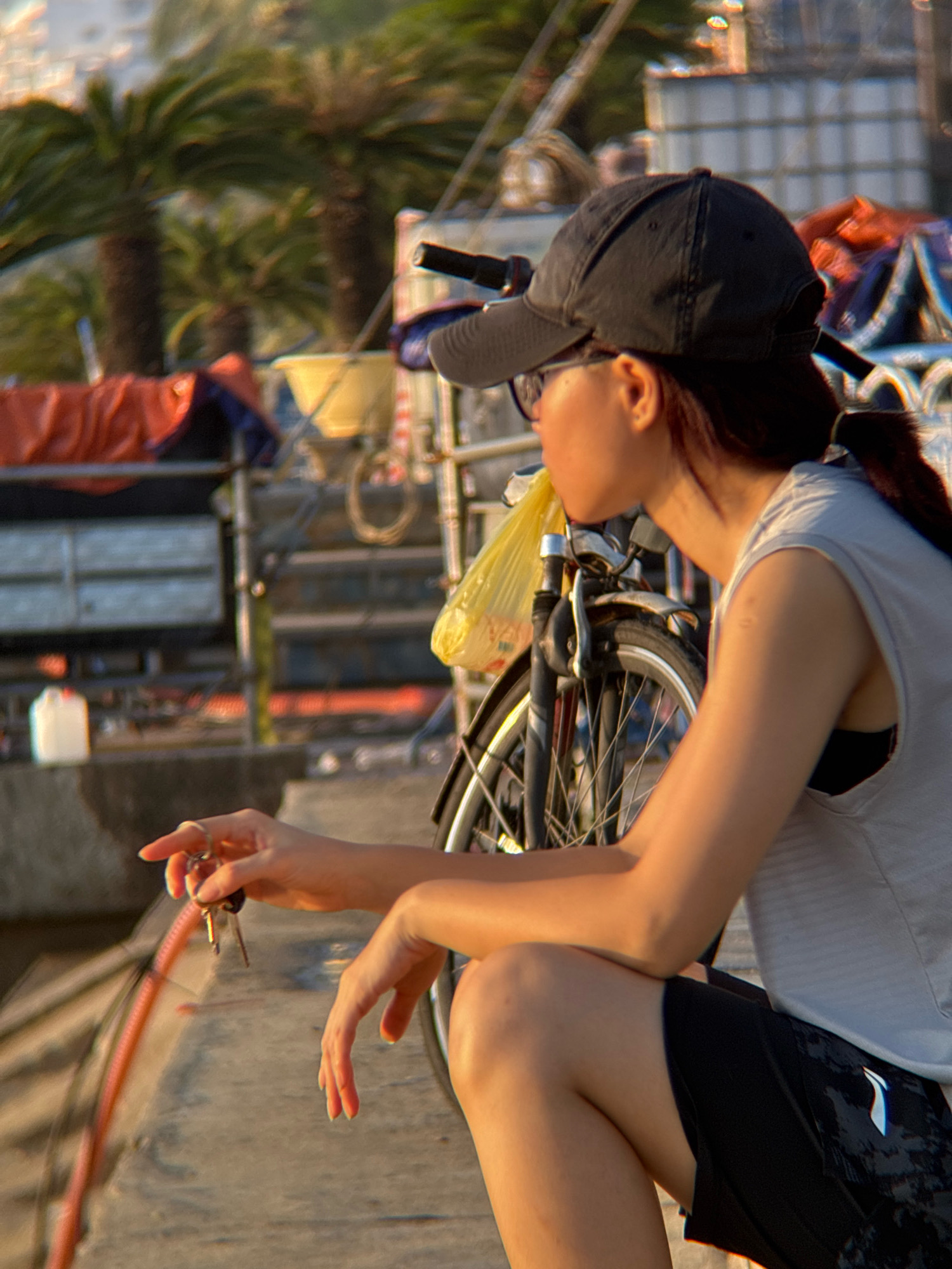 The image depicts a young Vietnamese woman with dark hair tied in a ponytail, wearing a black baseball cap, glasses, a light gray sleeveless top over a black sports bra, and black patterned shorts. She is holding a set of keys and a pink rope in her right hand, which is positioned in the foreground. Her left hand rests on her knee. She is seated next to a bicycle with a yellow plastic bag draped over its handlebars. The background shows lush palm trees, indicating a tropical or subtropical environment. The scene appears to be a waterfront area, possibly a harbor or pier, given the presence of boats and industrial-looking structures. The lighting suggests it is either early morning or late afternoon, with warm, golden sunlight illuminating the woman and her surroundings. The overall impression is one of casual relaxation in a scenic coastal setting. This scene is consistent with the environment of Nha Trang, Vietnam, known for its beaches, palm-lined boulevards, and active harbor.