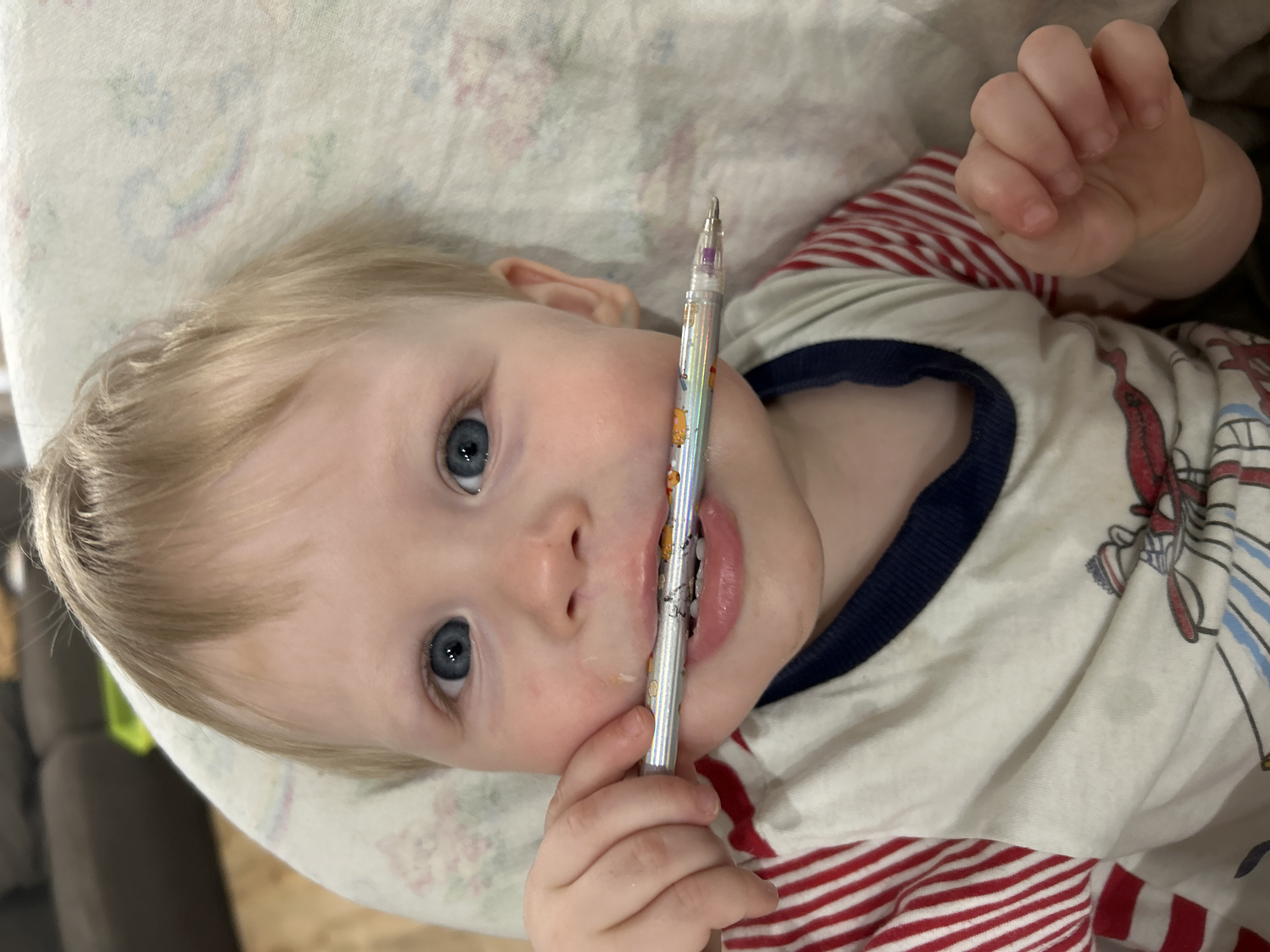 A young child with blond hair and blue eyes is lying down and holding a mechanical pencil in their mouth. The child is wearing a shirt with red and white stripes and a blue collar. The child is lying on a pillow or blanket with a light floral pattern. The location cannot be determined with certainty, as the image does not contain any visual cues or metadata that would provide information about the location, including whether it is in Chaikovsky, Russia.