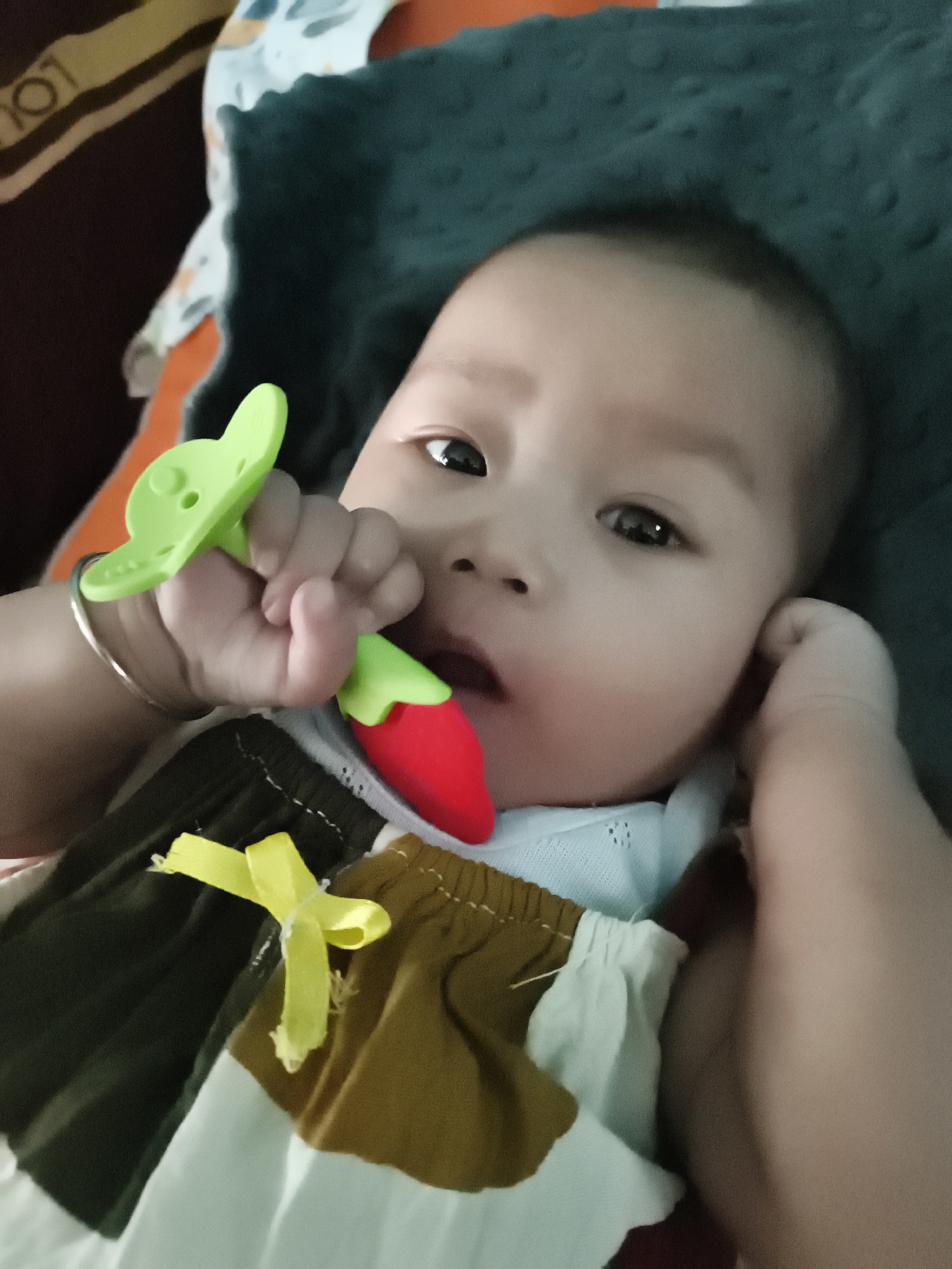 A close-up, slightly blurry, overhead shot shows a baby lying down and holding a red and green chili-shaped pacifier. The baby's right hand, adorned with a thin silver bracelet, is gripping the pacifier. The baby is wearing a white shirt with a blue collar and a multi-colored garment featuring blocks of white, dark green, and brown, accented with a yellow bow on the front. The baby's face is turned slightly to the right, with dark eyes looking towards the camera. The background consists of a dark green textured blanket. The image's quality suggests it was taken indoors in the city of Barengkrajan, Indonesia, likely in a residential setting.