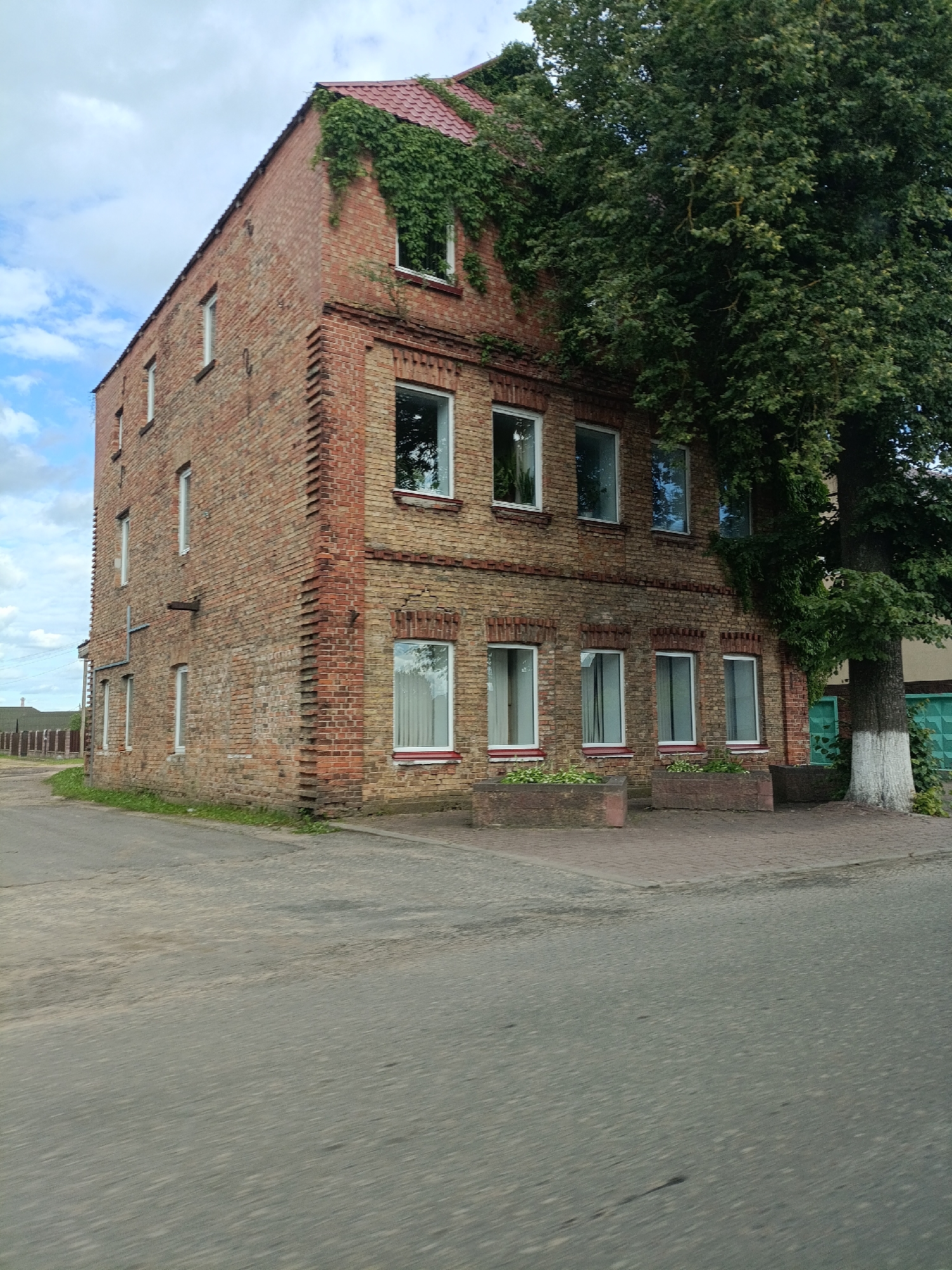 An exterior, eye-level, full shot shows a brick building overgrown with ivy. The building has a red, tiled roof and several windows. A large tree with dense green foliage stands next to the building on the right. The building is situated on a paved area in front of a sidewalk, which is made of bricks. Beyond the sidewalk is a patch of grass and then a road that curves away from the building. In the background, there is a wooden fence and some greenery. The sky is cloudy. The scene is set in a rural or suburban area, with no people visible.