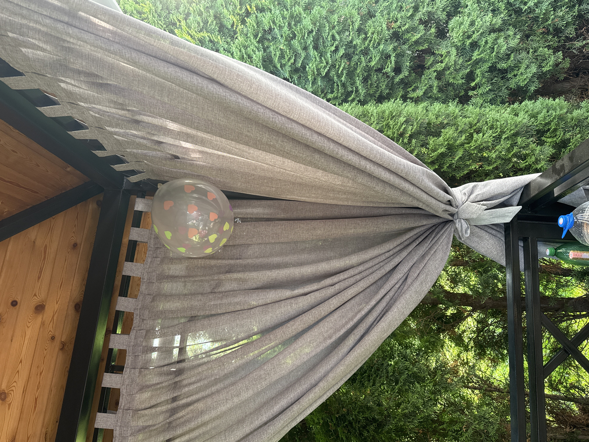 The image shows a clear balloon with heart-shaped confetti inside, hanging from a gray curtain tied to the side of a gazebo. The gazebo has a wooden ceiling and black metal supports. Outside the gazebo, there are green trees visible. The image appears to have been taken in Sary-Oy, Kyrgyzstan, although this is not explicitly shown in the image itself. The scene is outdoors and appears to be a bright, sunny day.