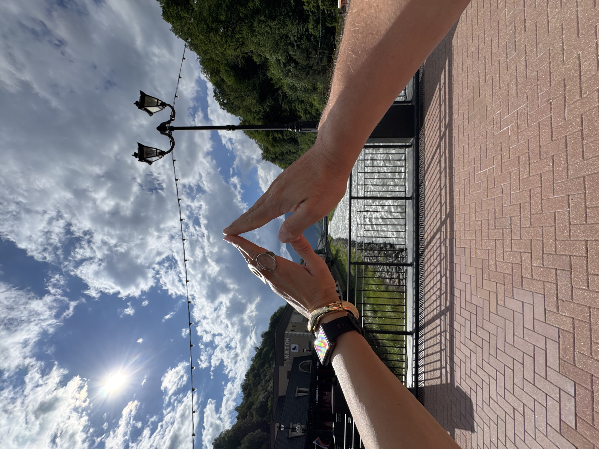 In Estosadok, Russia, two hands form an inverted triangle against a backdrop of a bright, cloudy sky, a river, and lush green mountains. The left hand, adorned with a smartwatch and several bracelets, is angled upwards, while the right hand is positioned similarly, with both hands meeting at the fingertips to create the triangular shape. In the middle ground, a river flows swiftly, bordered by a black metal railing. Beyond the river, buildings with "КАТОК" signage are visible, nestled at the foot of a densely forested mountain. A streetlamp with two lights stands prominently near the center of the frame, and a string of decorative lights stretches across the sky. The foreground is paved with reddish-brown interlocking bricks laid in a herringbone pattern, with shadows cast by the railing and the hands.