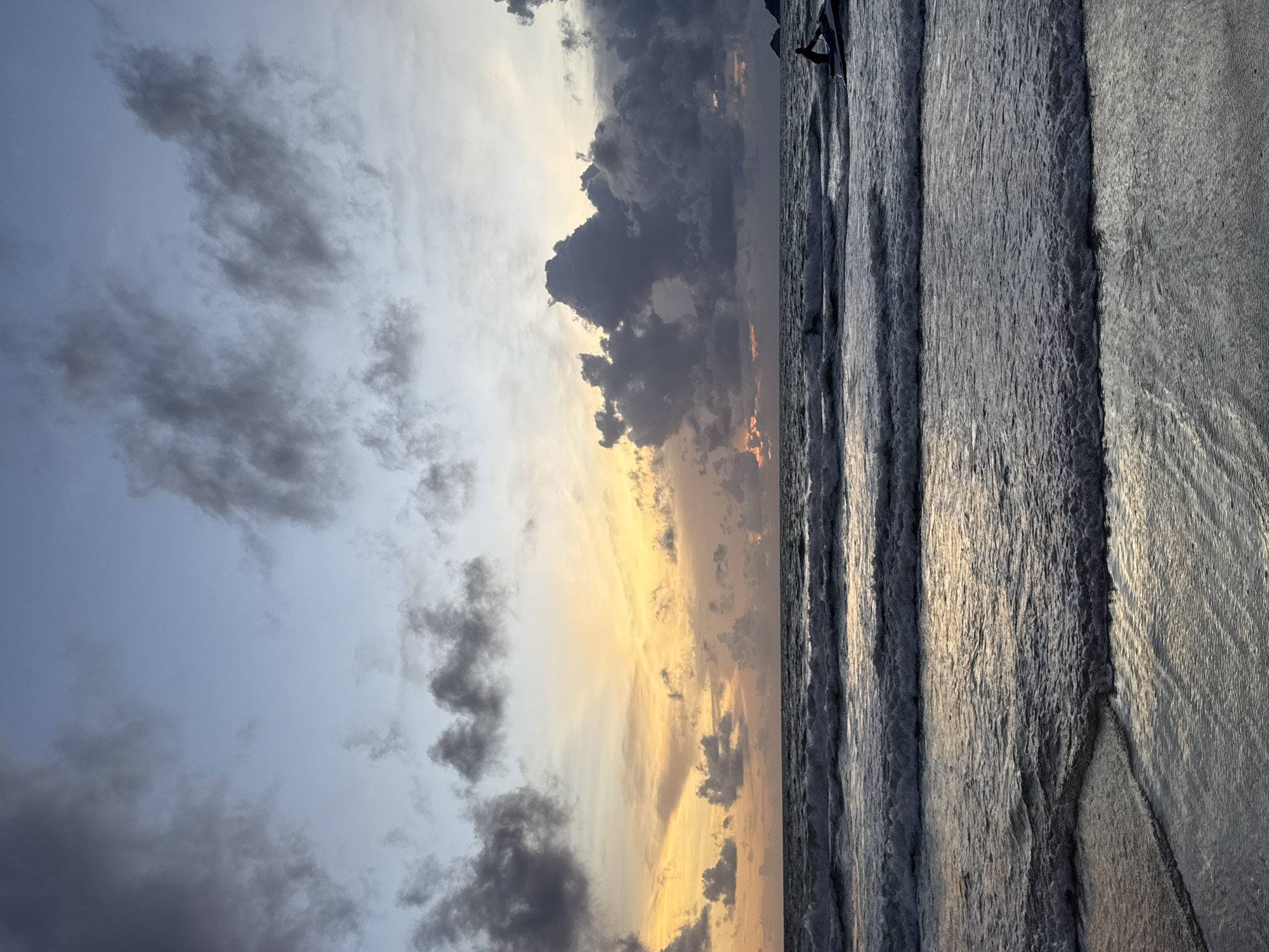 The image depicts a sunset scene at Patong Beach, Thailand. The sky is a gradient of blues and yellows, with dramatic dark clouds scattered across the horizon. Waves are crashing on the shore, creating a frothy white surf that recedes over the wet sand. In the distance, a person is riding a jet ski, silhouetted against the water. The overall atmosphere is one of twilight calm, with the last light of day illuminating the ocean and sky.