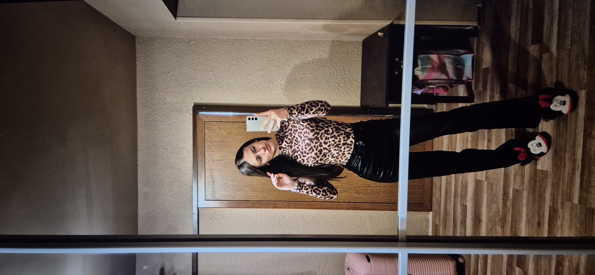A young woman in a leopard-print top and black pants stands in what appears to be a room with wood-patterned flooring, taking a selfie in a mirror. She is holding a smartphone with a three-camera setup. The room features a brown door, a dark cabinet or shelf unit with items inside, and a pink suitcase partially visible at the bottom of the mirror's reflection. The woman wears black slippers adorned with Mickey Mouse faces. The background shows textured walls and a muted color palette. The image does not provide enough information to specifically locate the room in Sofia, Bulgaria, or to describe any actions or interactions outside of the woman taking a selfie.
