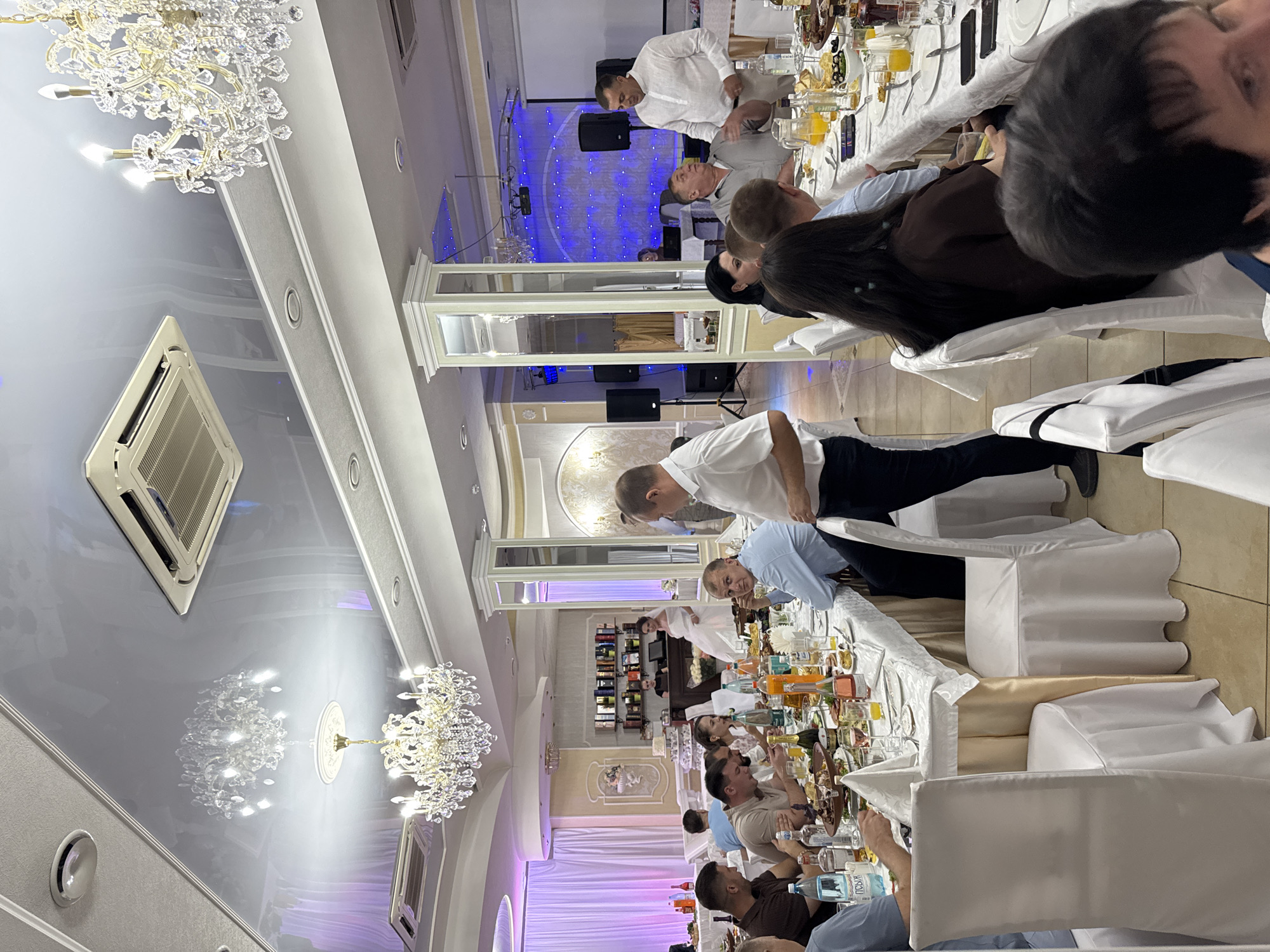 The image depicts an indoor banquet hall, likely a wedding reception, set within a building located in Ust-Labinsk, Russia. The hall is elegantly decorated with chandeliers and ambient lighting. Several tables are laden with food and drinks, indicating a celebratory meal. Guests, dressed in formal attire, are seated at the tables and mingling. Some individuals are engaged in conversation, while others are attending to the food. A bride, wearing a white wedding dress, is visible in the background, standing near a cake display. The overall scene suggests a lively and festive gathering.