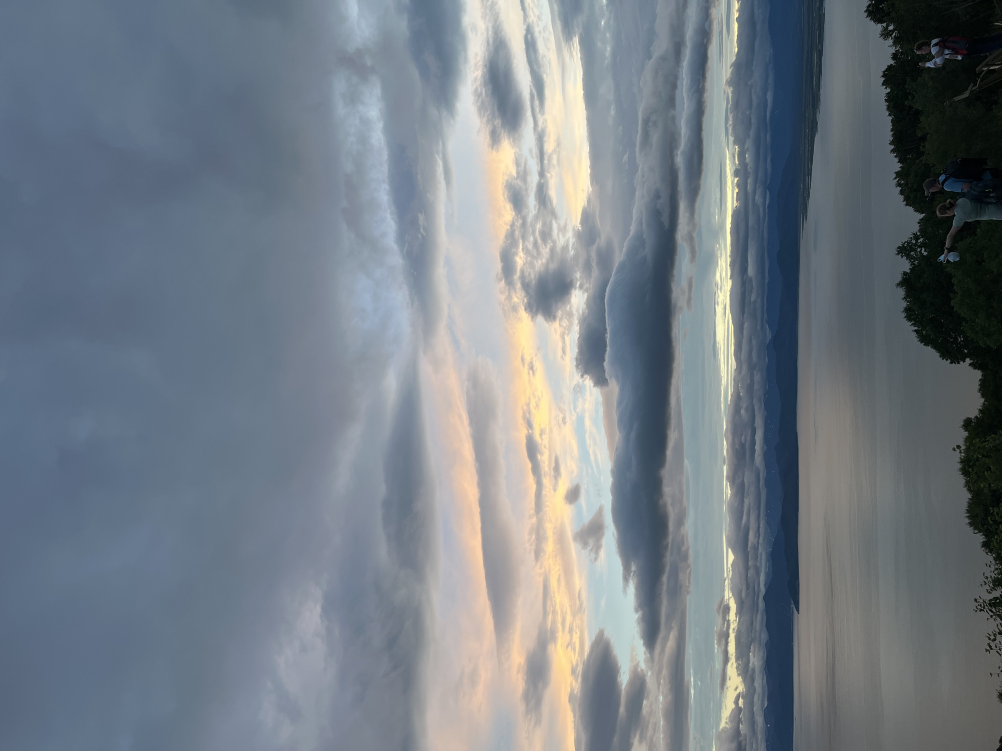 The image captures a panoramic view of a large body of water, likely Avacha Bay, under a dramatic sky filled with layered clouds. The water's surface is calm, reflecting the muted colors of the sky, which range from deep grays and blues to soft pinks and yellows near the horizon. In the distance, a mountain range forms a dark silhouette against the sky. In the foreground, a lush green treeline partially obscures a group of people.
Three individuals are visible at the edge of the treeline, looking out towards the water and mountains. On the left, a woman in a light-colored top and dark pants holds a white object, possibly a phone or camera, in her raised right hand. She is looking to her left. Beside her, a man wearing a dark blue t-shirt and a backpack is also looking in the same direction as the woman. To the right of the couple, a third person, wearing a white top and a backpack, is holding a phone up to take a picture or video. The group appears to be engaged in observing the landscape. The overall impression is that of a scenic overlook, possibly near Petropavlovsk-Kamchatsky, Russia, given the geography.
