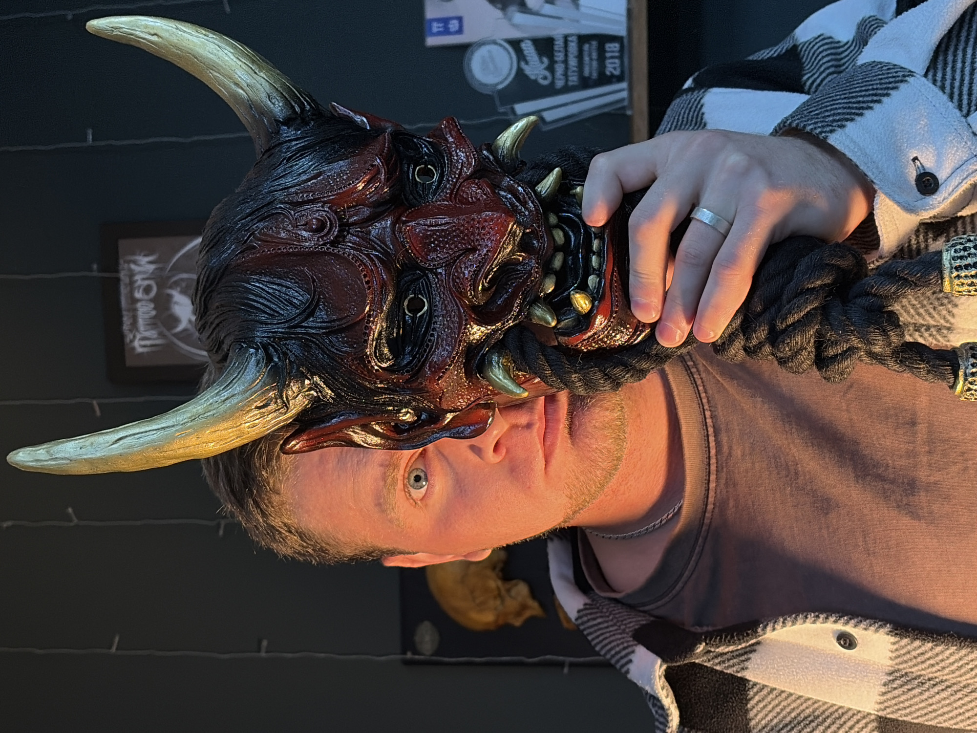A close-up portrait shows a light-skinned man with short, light brown hair and a closely trimmed beard. He is wearing a grey t-shirt under a black and white plaid shirt. His right hand, adorned with a silver ring, rests on a black rope tied to a large, intricately detailed, red and black Oni mask with prominent gold horns. The mask dominates the upper part of the frame.
In the background, there's a framed "Tattoo OK" logo and what appears to be a black shelf with various items on it. Lower down, part of a skull-shaped object is visible.
The location is presumed to be Chaikovsky, Russia, based on potential metadata associated with the image. However, this detail is not directly apparent within the image itself.