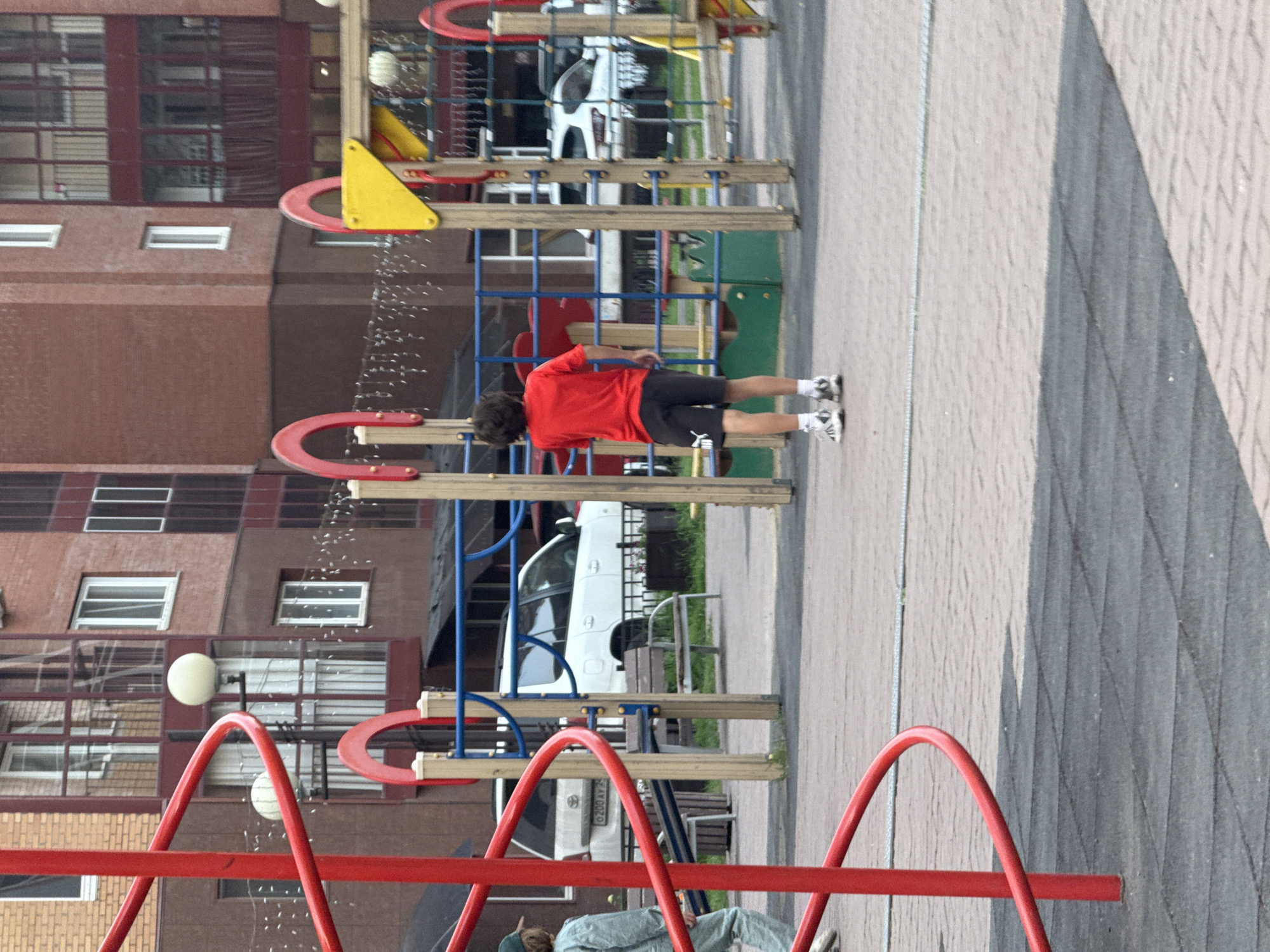 In Krasnoyarsk, Russia, a young person in a red t-shirt and black shorts is exercising on a playground. The playground equipment is made of metal and wood, with red and blue accents. There are several structures, including what appears to be a climbing frame and a swing set. In the background, a multi-story apartment building is visible, with rows of windows. The weather appears to be sunny and pleasant. The person is in mid-motion, suggesting an active and energetic moment. The overall scene depicts a common urban recreational space.