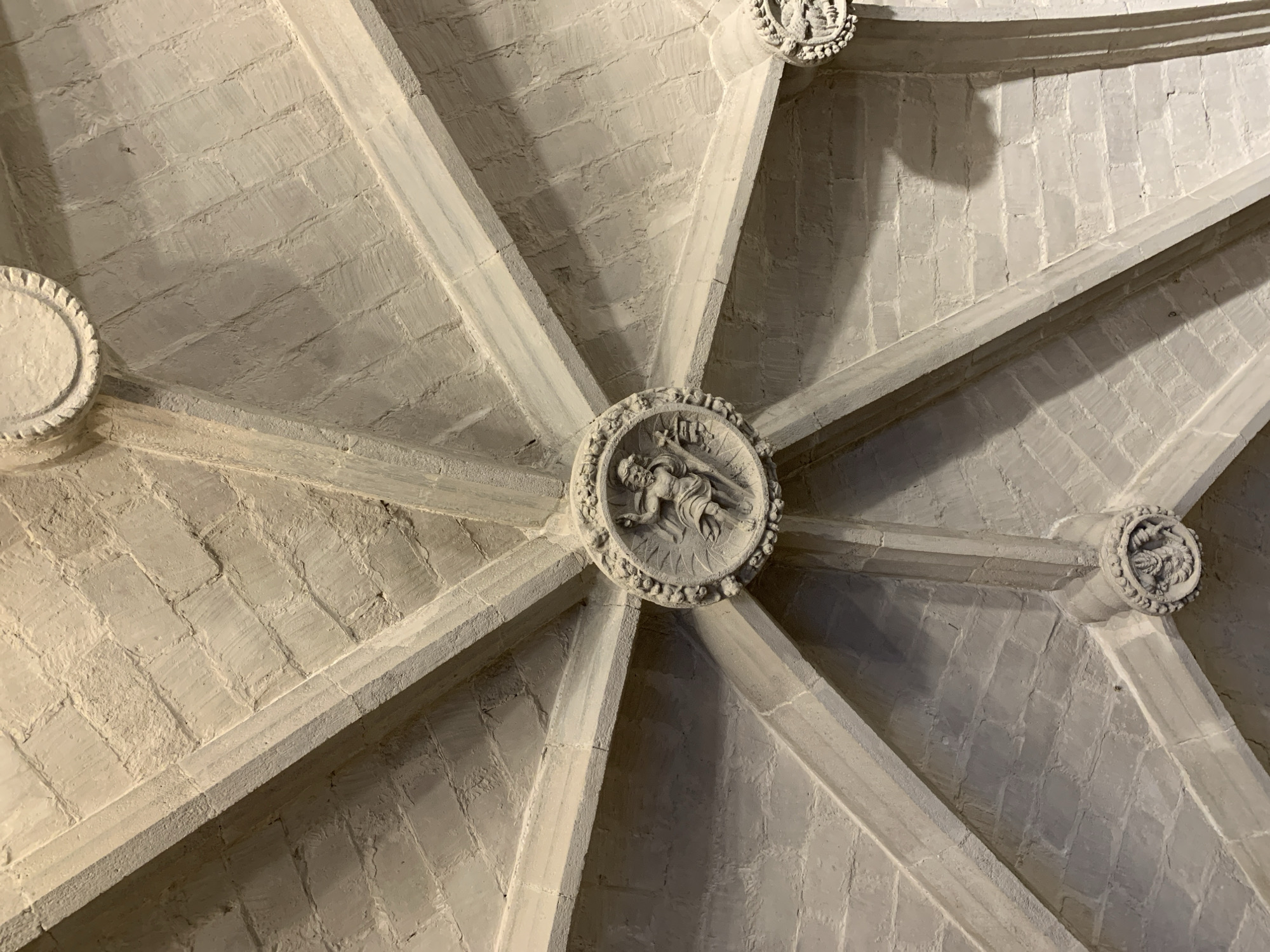This is a detailed view of the ceiling of a gothic-style building, specifically featuring the ribbed vaulting, a common architectural element in such structures. The central boss of the vault is adorned with a circular relief depicting a human figure in motion. The surrounding ribs, constructed from stone, converge at this point, and additional decorative bosses are visible at other intersections. The structure suggests an old, possibly religious building. As for its location in Barcelona, Spain, without further information, it is difficult to pinpoint which specific site this vault belongs to, although the gothic style suggests it could be a church or cathedral, as Barcelona is well-known for such architecture, including the Barcelona Cathedral (also known as the Cathedral of Santa Eulalia) or the Basilica de Santa Maria del Mar.