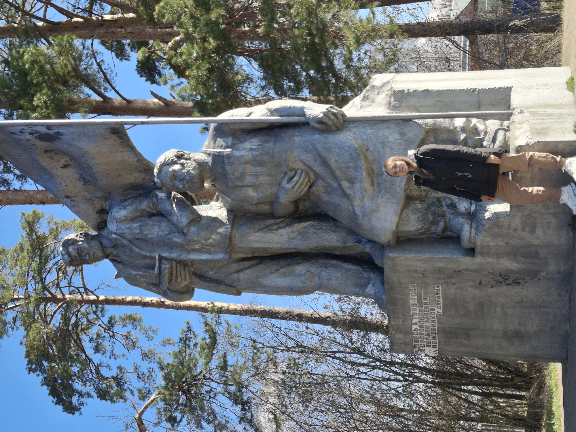 The image captures a monument in Sarapul, Russia, featuring two Soviet-era figures rendered in stone. The figures appear to be holding a banner or flag. The monument has a concrete base with Cyrillic inscriptions. A person stands next to the monument on the right side of the frame, posing for a photo. The background includes trees with visible branches and a clear blue sky above. The image is taken during daytime, suggesting a sunny environment. The monument seems to be located outdoors in a park or public space.