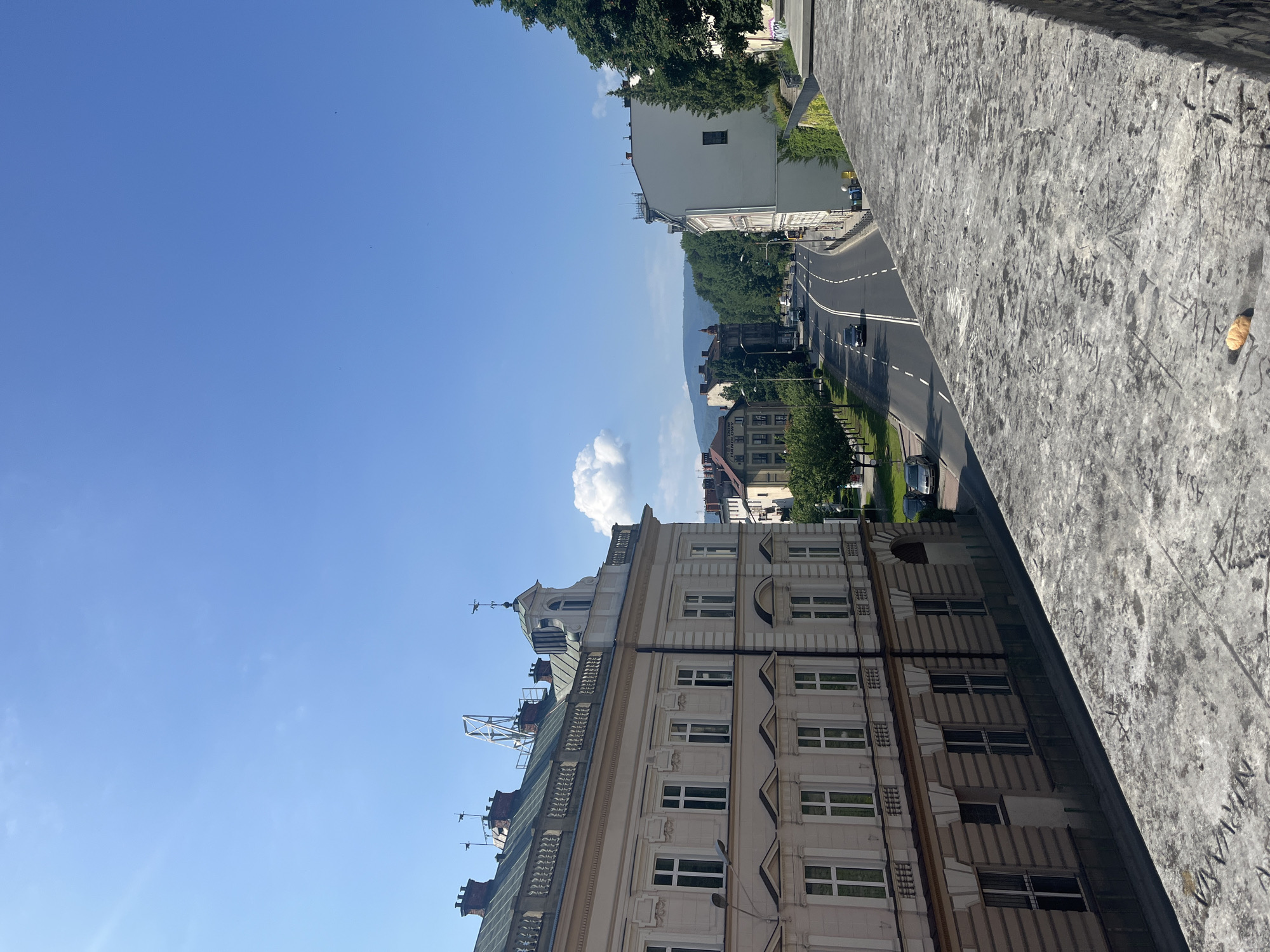 The image depicts a street scene in a city on a sunny day. In the foreground, a weathered stone wall is visible, inscribed with graffiti. Beyond the wall, a street curves to the right, lined with buildings on both sides. On the left, a multi-story building with a pale facade and numerous windows is prominent. Across the street, more buildings and lush green trees line the road. Cars are driving on the street, and the sky is a clear blue with a few scattered clouds. In the distance, hills or mountains are visible.