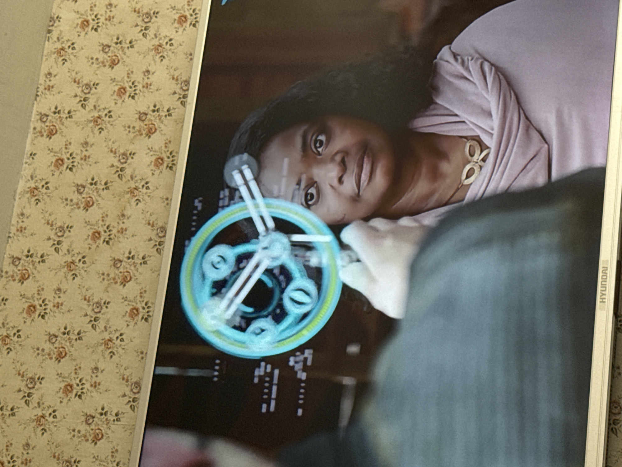 The image shows a Hyundai monitor displaying a scene with a dark-skinned woman. She is looking directly at the viewer with a slight smile, while a blue holographic circle with inner shapes is overlaid on the screen. The woman is wearing a light purple top and a gold necklace with a butterfly or bow-shaped pendant. Part of another person's shoulder, covered in gray fabric, is visible in the foreground, partially obscuring the lower right part of the screen. The background behind the monitor shows a floral-patterned surface. The image does not contain information about Mytishchi, Russia.