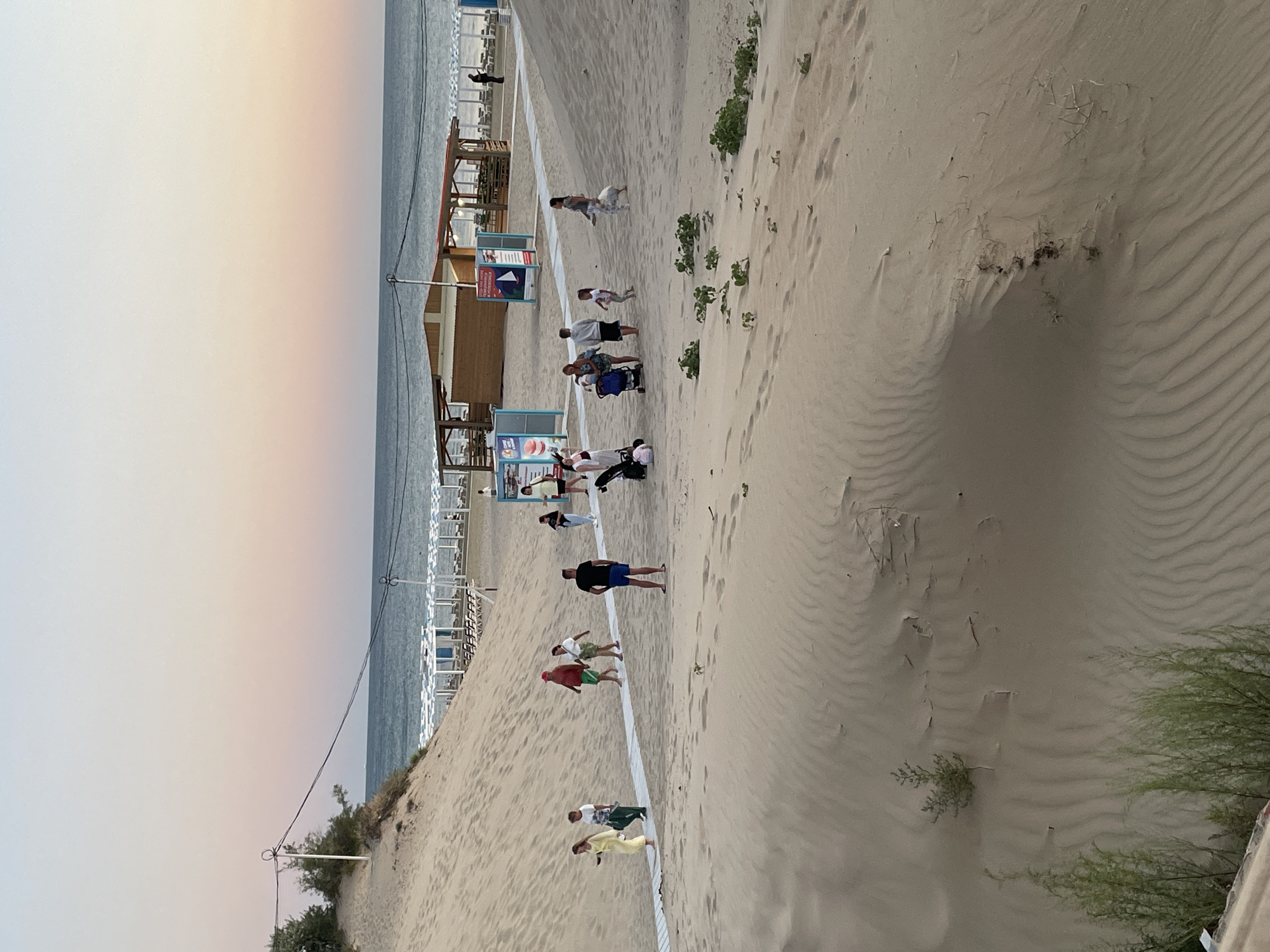 The image depicts a sandy beach scene with a group of people walking and relaxing. The sky is a soft gradient of pink and blue, suggesting either sunrise or sunset. In the foreground, sand dunes with rippled textures are visible, with some sparse vegetation growing on them. A wooden walkway runs across the sand, with several individuals walking on it.
Further back, a beach cafe or shelter with a wooden structure and a red roof is situated. Beach chairs and umbrellas are set up on the sand beyond the cafe, facing the ocean. The ocean is calm and appears to stretch to the horizon. Several people are seen in various groups: some are walking individually or in pairs, while others appear to be with children or pushing strollers. One person is standing near the cafe, while others are scattered across the beach. Power lines are visible in the upper portion of the image, strung between poles. The overall atmosphere suggests a peaceful, end-of-day scene at a beach resort.