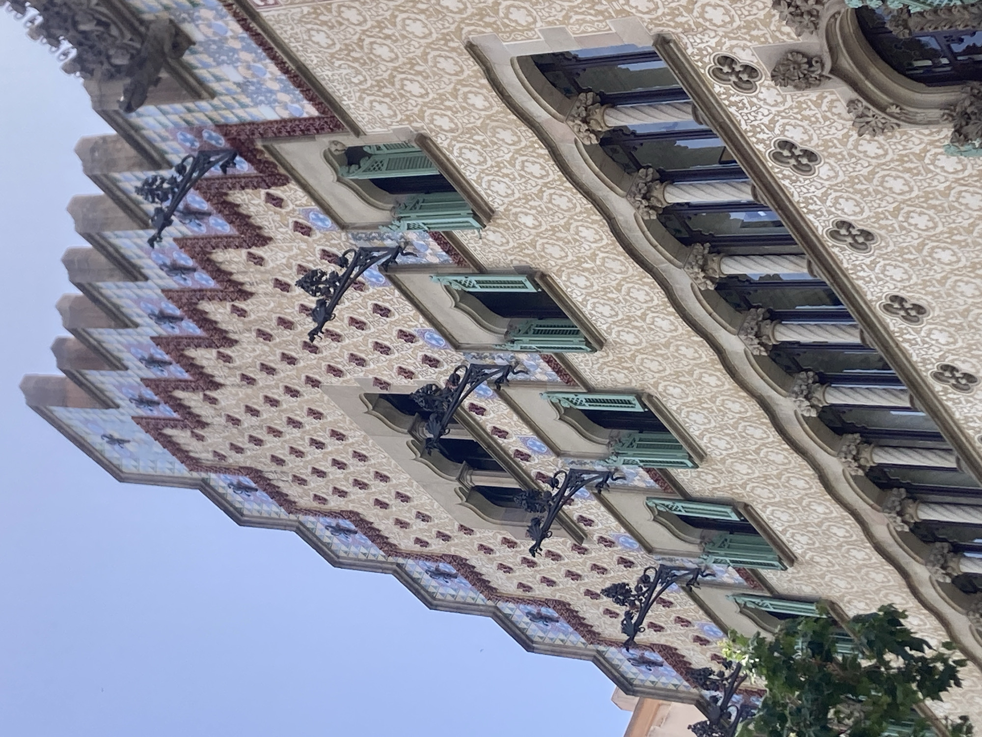 The image shows the facade of Casa Batlló, a building designed by Antoni Gaudí, located on Passeig de Gràcia in the Eixample district of Barcelona, Spain. The facade is characterized by its organic, undulating forms and vibrant, colorful ornamentation.
The upper part of the building features a stepped gable, decorated with ceramic tiles in various geometric patterns and colors, including blues, whites, and reds. Interspersed among the tiles are dark, wrought-iron sconces, designed to resemble fantastical creatures or floral motifs, projecting outwards.
Below the gable, several windows are visible, each with a distinct shape and surrounded by decorative stonework. Some of these windows are partially covered by mint-green shutters. The main body of the facade is covered in a textured plasterwork adorned with a repeating pattern of stylized floral or organic shapes in white and beige.
The lower section of the facade, visible towards the bottom of the image, features arched windows supported by decorative columns with twisted shafts and intricately carved capitals. The stonework around these lower windows also incorporates a repeating decorative motif. The sky above is clear and blue, indicating a daytime scene. A small portion of a tree with green leaves is visible on the left side of the image, suggesting the building is situated on a street with greenery. No people are visible in the image.