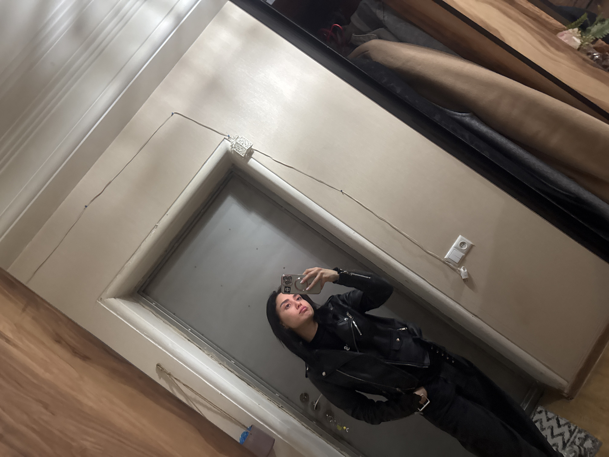 A woman stands in front of a mirror, taking a selfie. She is wearing a black leather jacket and black pants. The mirror is framed in a light-colored wood and the surrounding wall is a neutral beige. A light switch is visible on the wall. The floor is a light-colored wood laminate, and a small rug is partially visible in the corner. The location is unknown, and there is no information indicating it is Tbilisi, Georgia.