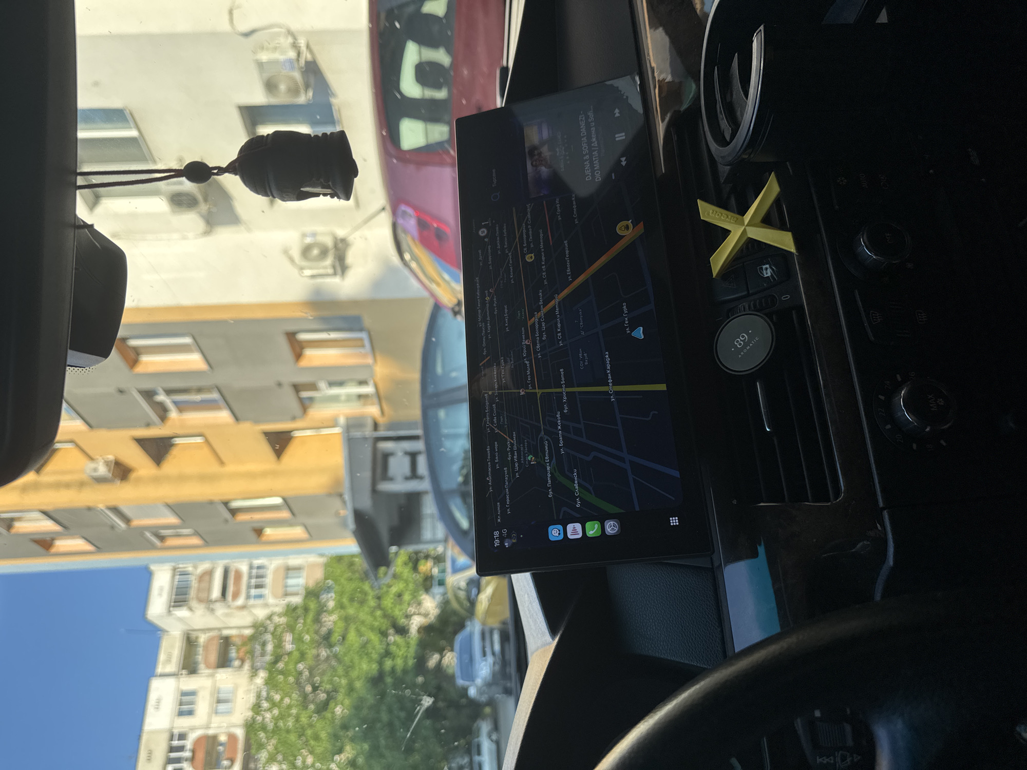 The image depicts the interior of a vehicle, specifically a BMW 5 Series (E60/E61 generation), viewed from the driver's or front passenger's perspective, looking forward through the windshield into an urban environment.
The dashboard, dark in color with wood trim accents, occupies the lower portion of the frame. The left side of a black steering wheel is visible on the far left. A large, rectangular infotainment screen is prominently mounted centrally on the dashboard, displaying a map application. The map shows street names in Cyrillic, identifying the location as Stara Zagora, Bulgaria, with prominent streets such as "бул. Патриарх Евтимий" and "бул. Славянски" visible. The screen indicates a time of 19:18 and a 4G cellular signal. App icons for phone, messages, and a music player are visible on the left side of the screen. A music widget on the right displays "DJENA & SOFIA DANEZI - DIO MATIA".
To the right of the screen, air vents are present, with a circular black air freshener labeled ".89 AROMATIC" in the leftmost vent and a yellow 'X'-shaped air freshener in the adjacent vent. Below the vents, climate control dials are partially visible, showing temperature settings from 18 to 26 degrees Celsius and a "MAX" setting. A black cup holder is visible on the far right.
Attached to the rearview mirror support, a dark, bell-shaped ornament hangs centrally at the top of the windshield.
Outside the vehicle, the environment is an urban street under clear, blue skies, illuminated by direct sunlight, consistent with the 19:18 timestamp in late afternoon or early evening. A multi-story yellow/orange residential building with numerous windows and air conditioning units dominates the right side of the view. Behind it, a lighter-colored, similar residential building is visible. Green trees are interspersed between the buildings, primarily on the left. Several parked or slowly moving vehicles are visible: a silver sedan/SUV on the left, a yellow car further back, and a red car (possibly a Fiat Punto) in the foreground behind the infotainment screen. No individuals are visible in the scene.