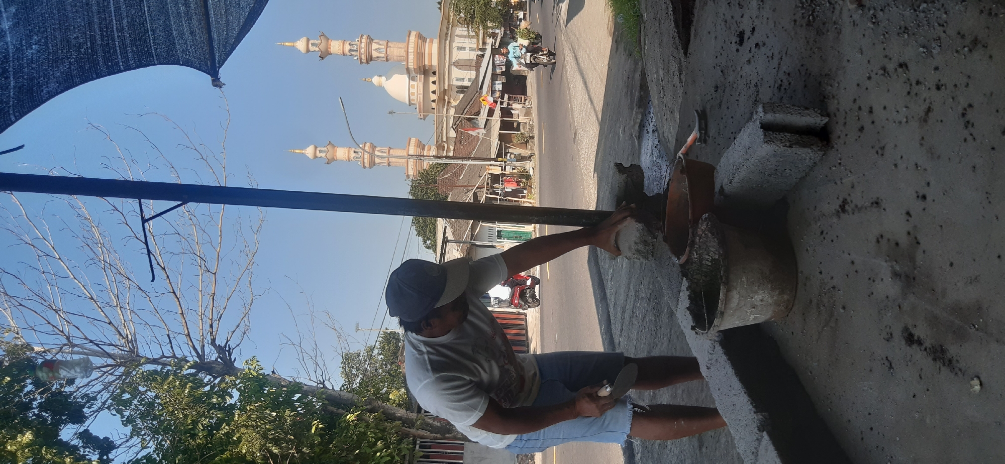 A man wearing a white t-shirt and blue shorts is working on a construction project in Sepakek, Indonesia. He is holding a trowel in his left hand and using his right hand to place a piece of concrete. In front of him are two buckets, one filled with concrete mix and the other an empty orange basin. The scene takes place outdoors, with a street and buildings in the background, including a mosque with two minarets. A motorcycle is parked nearby, and another motorcycle is visible on the street. The sky is clear and blue.