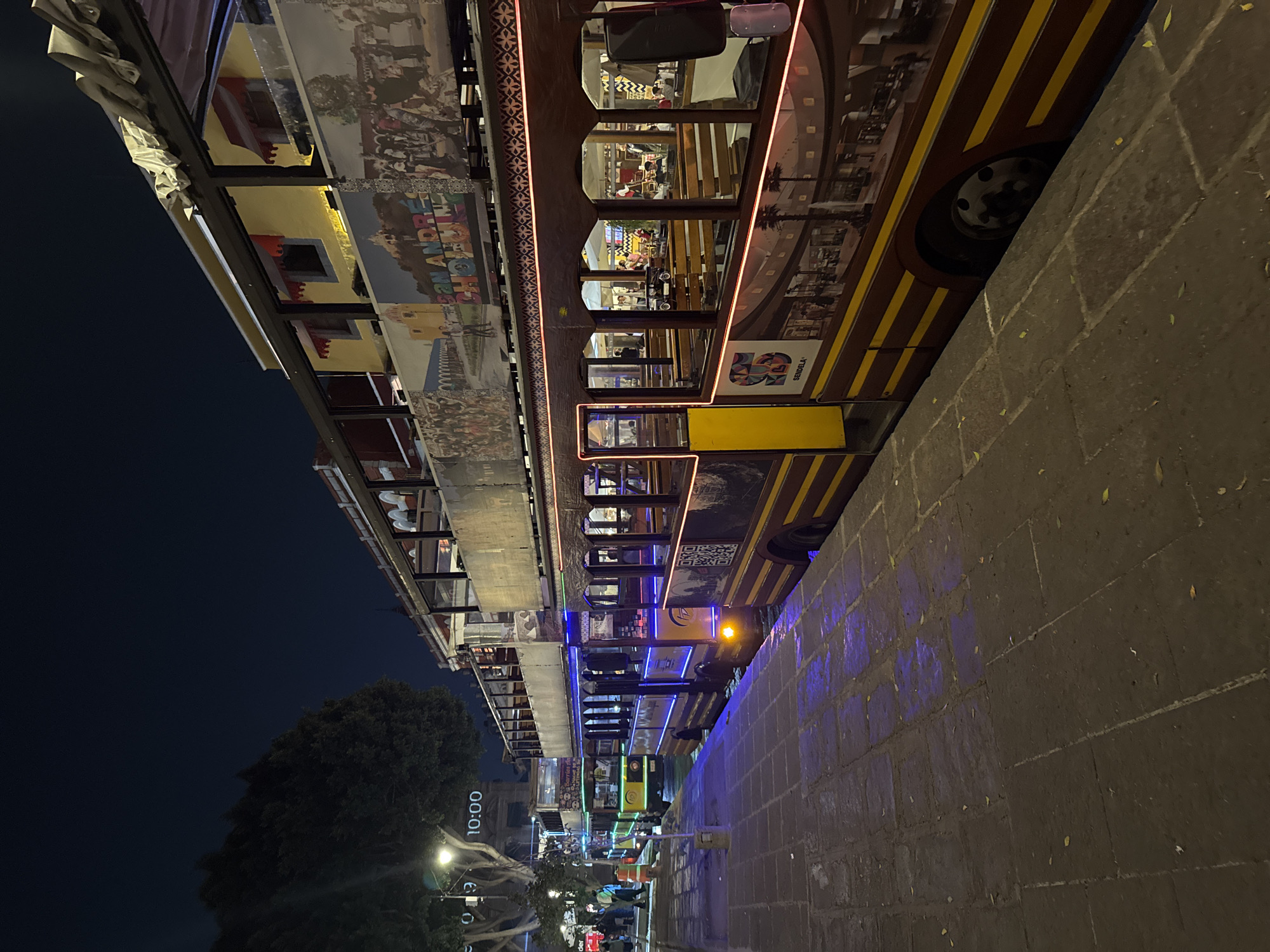 At night, an illuminated double-decker tourist bus passes along a brick-paved sidewalk in Puebla, Mexico. The bus has brown and yellow stripes and windows through which people can be seen. The bus exterior is decorated with images of historical scenes and local landmarks, one of which reads "San Andrés Cholula." Blue lights illuminate the sidewalk next to the bus. In the distance, streetlights illuminate the street and pedestrians on the sidewalk. A large tree and a building with a clock tower are visible. The sky is dark.