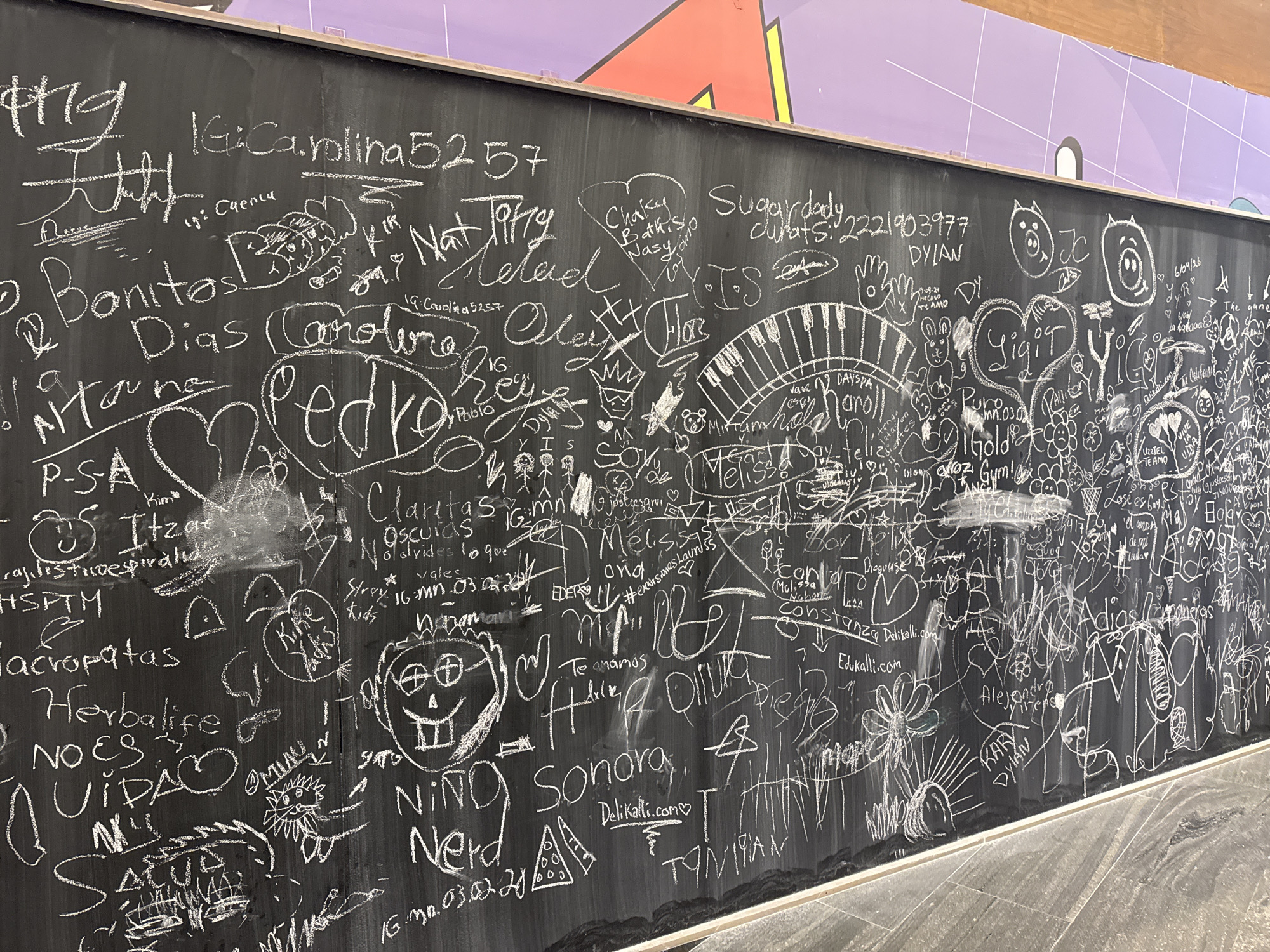 The image shows a chalkboard wall covered in white chalk graffiti. The writing is dense and overlapping, with names, dates, phrases, and drawings. Some of the legible words include "Bonitos Dias," "Carolina," "Pedro," "Herbalife," "Sonora," and "Adios." There are also drawings of faces, hearts, a piano keyboard, and various other symbols. The context of the graffiti suggests a public space where people have left their mark. Some text points to the Delikalli Ice cream shop, located in the city of Puebla, Mexico. The presence of dates indicates that the graffiti has accumulated over time. The bottom of the image shows the wall meeting the floor, suggesting that it is an interior space.