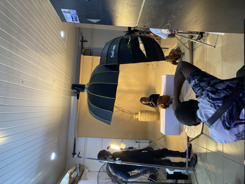 The image depicts an active photography studio located in Bukuru, Nigeria. In the immediate foreground, a male individual, observed from behind, is positioned over a circular silver reflector on the tiled floor, holding a camera pointed towards the subjects. To the left, a second male individual, dressed in a dark t-shirt and jeans, stands near a metal light stand equipped with a white fan and two square LED panel lights.
A seamless light brown backdrop occupies the central area of the studio. On a white rectangular prop situated on this backdrop, a young girl with dark braided hair, wearing a black top and a patterned orange and black skirt, sits, looking towards the camera. Behind her, a tall, woven vase containing several bare branches stands as a decorative element. To the right of the girl, a large black photography umbrella softbox, bearing the "Selens" brand name, is mounted on a light stand, directing light towards the scene. Partially visible beneath this softbox, a toddler in a patterned outfit crawls on a low wooden platform on the floor. The studio ceiling is constructed from white slat panels, featuring several recessed circular lights. A dark wooden panel wall with a white poster attached is visible on the far right side of the frame.