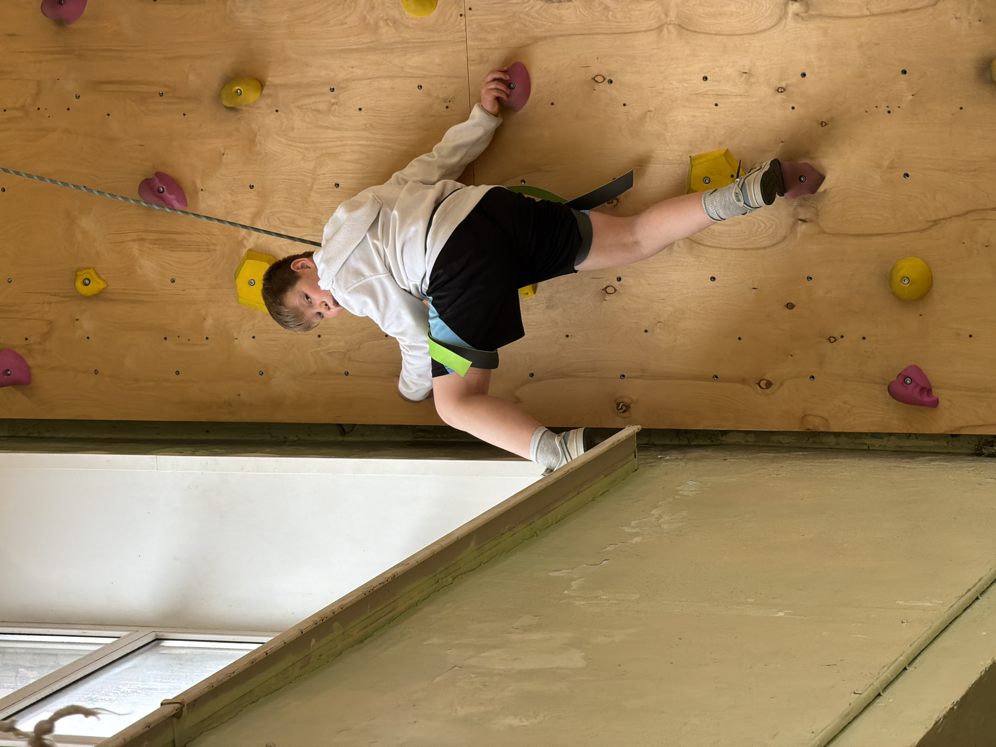 A young boy is depicted in mid-action on an indoor rock climbing wall. He is positioned horizontally across the frame, actively engaged in climbing or traversing. The boy wears a white long-sleeved hooded top, black shorts with light blue accents, light-colored socks, and what appear to be climbing shoes or sneakers. A black and green climbing harness is visible around his waist, connected to a grey and green speckled climbing rope that stretches across the wooden wall.
He holds onto a pink climbing hold with his right hand, while his left foot rests on another pink hold. His right foot is placed on a yellow hold. The boy is looking towards the camera with a neutral expression.
The climbing wall itself is constructed from light-toned wooden panels, fitted with numerous brightly colored climbing holds in yellow and pink, along with visible screw holes. Below the climbing wall, a portion of a white surface, possibly a wall or ceiling, is visible, along with a window or door frame. Further down, a light green-painted surface, resembling a floor or a lower structural element, shows signs of wear.
The setting is an indoor climbing facility, likely a gym or recreational area, during daylight hours, given the natural light filtering in. The activity clearly shows rock climbing or bouldering, taking place in Izhevsk, Russia. There is no visible text in the image.