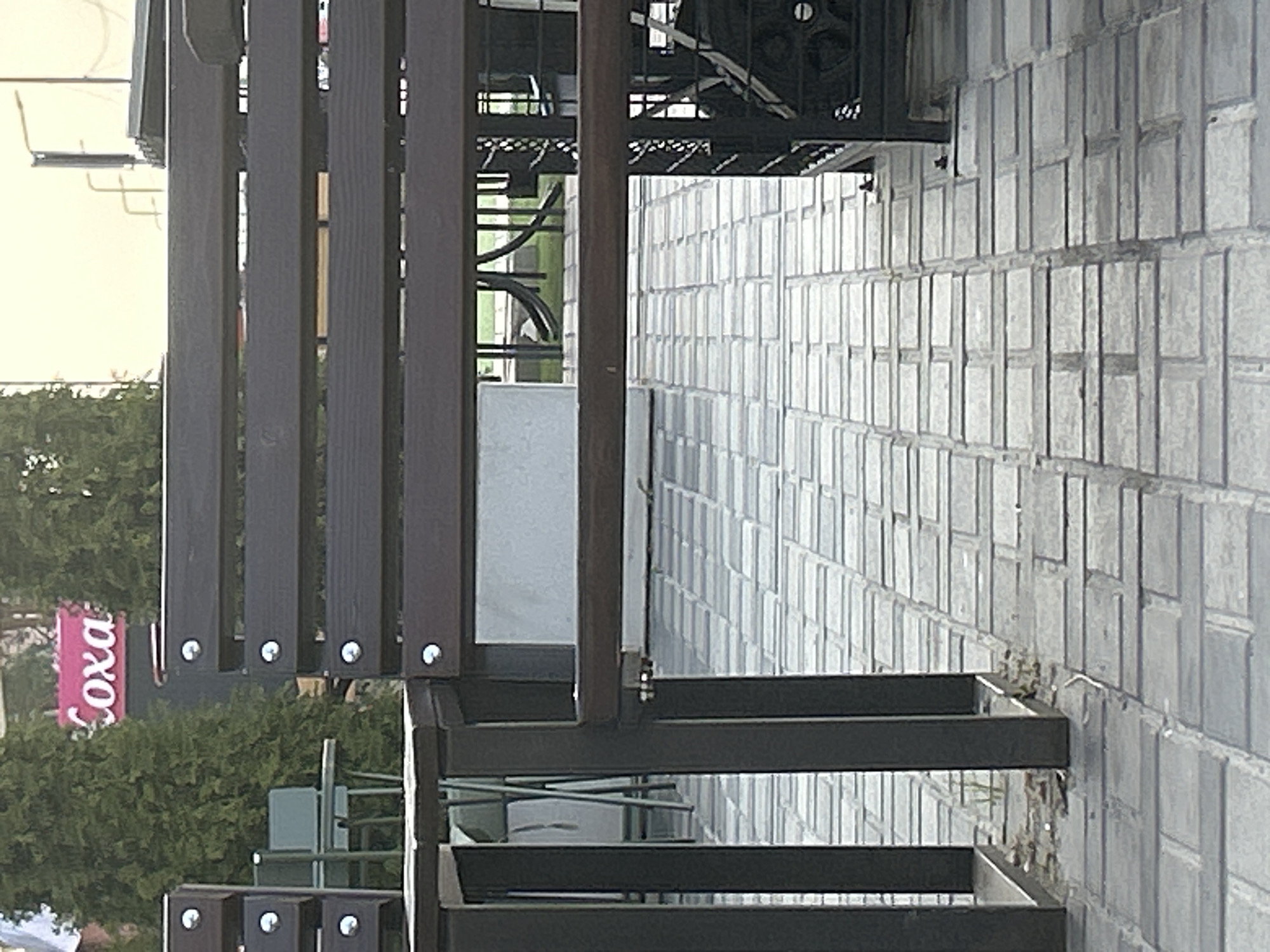 The image depicts an outdoor seating area with dark wooden benches and metal supports, situated on a paved patio. In the background, a pink sign with the text "Coxa" is visible, along with green trees and a light-colored building. A small table and chairs can be seen to the left of the benches, and more seating and architectural elements are present further back. The scene is located in Brovary, Ukraine.