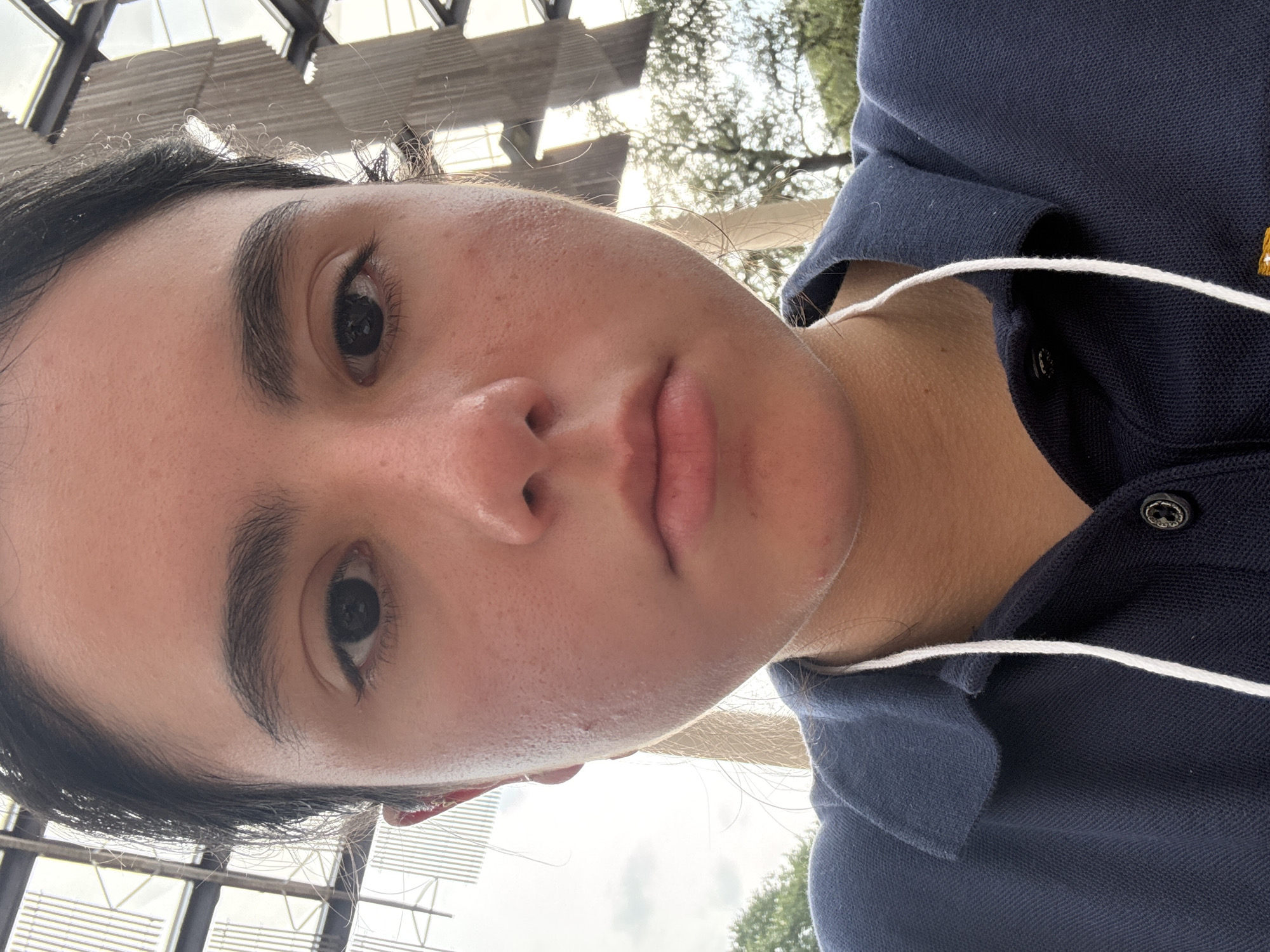 A close-up selfie of a person with dark hair and eyes, wearing a dark blue polo shirt with white trim, is taken from a low angle. The person has a neutral expression. White earbud cords are visible around the neck. The background shows a structural roof element of a building, and hints of trees and sky. The location is suggested to be San Antonio, United States of America.