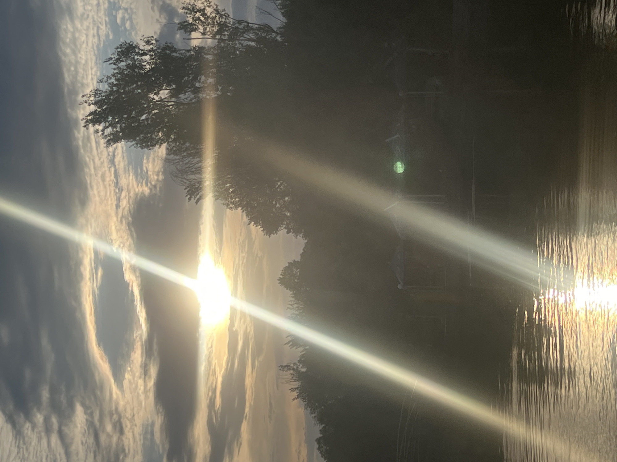 The image captures a tranquil scene of a body of water reflecting the sun's rays. The sun, a bright orb, is partially obscured by clouds, casting a golden glow across the sky and the water's surface. Streaks of light pierce through the atmosphere, creating a dramatic effect. Trees line the edge of the water, their dark silhouettes contrasting with the luminous sky. The overall atmosphere is serene and peaceful, suggesting a moment of natural beauty. The time of day appears to be late afternoon or early evening, given the warm, low-angled sunlight. The location is identified as Yerevan, Armenia. There is no visible text in the image.