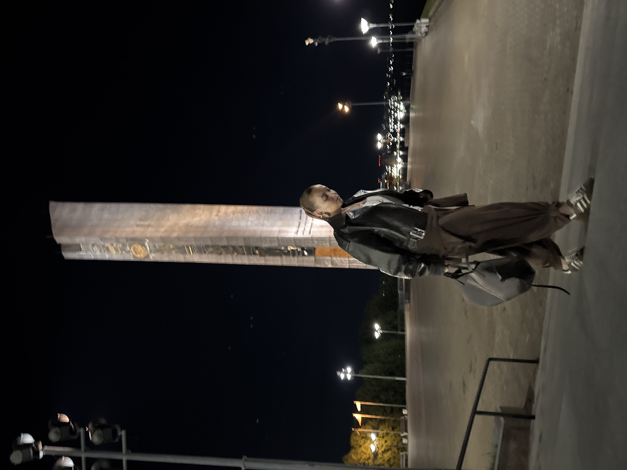 The image captures a woman standing near the monument "Forever with Russia", located in Izhevsk, Russia. The monument appears tall and is illuminated, contrasting against the dark night sky. The woman is wearing a leather jacket, brown pants, and sneakers, and she is carrying a gray bag. Streetlights are visible in the background, lining the path. The surroundings include trees and a paved area, indicating an urban setting.