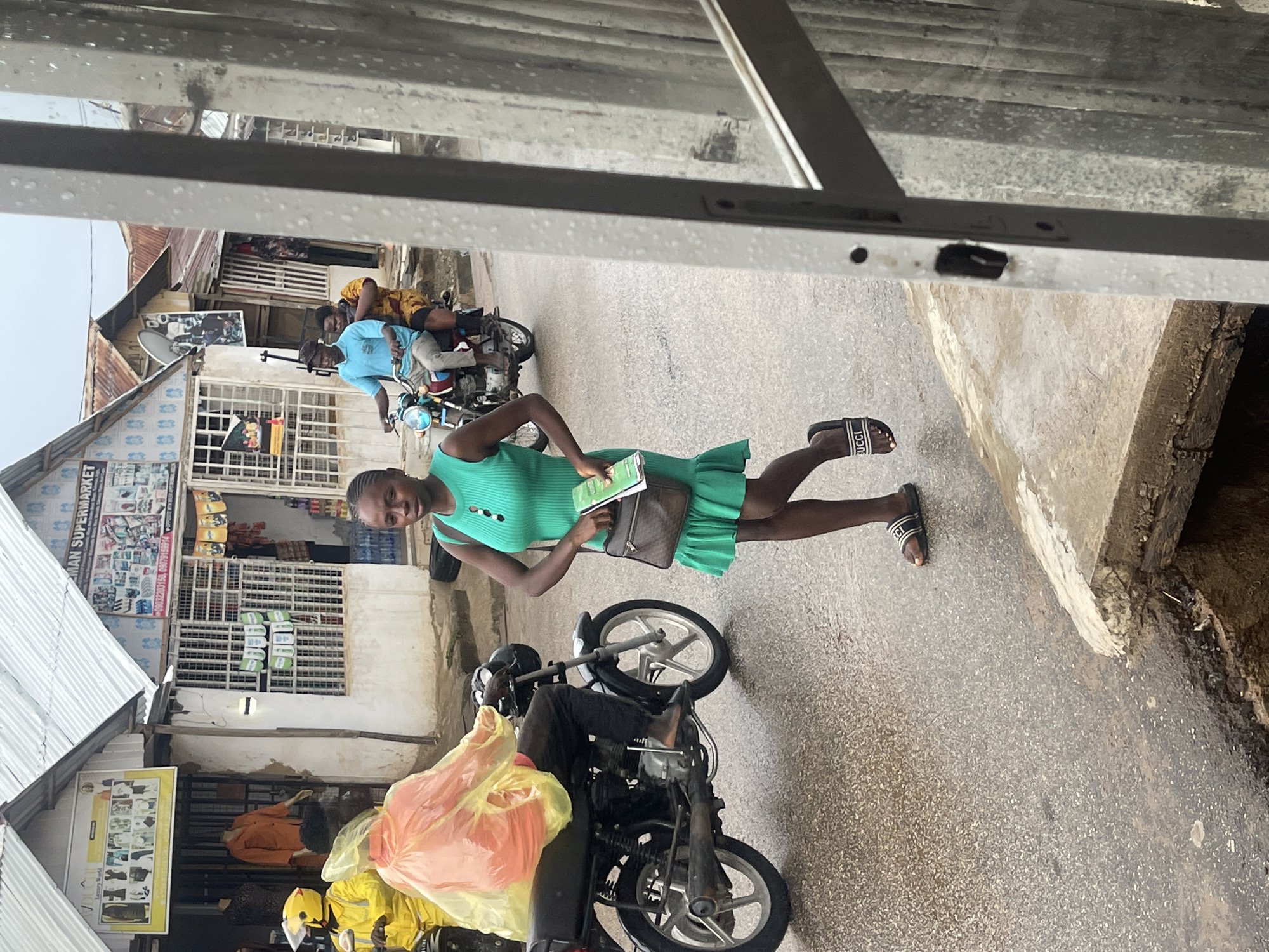 This image depicts a lively street scene in what appears to be a commercial or market area in Abuja, Nigeria. The central figure is a young woman, positioned towards the left of the frame, smiling brightly at the camera. She wears a sleeveless, teal green mini-dress and black slide sandals with "GUCCI" written on them. She holds a light-green notebook or book in her hands and has a small, dark shoulder bag.
The setting features various buildings with corrugated metal roofs and shopfronts lining a wet, asphalt road, suggesting recent rain or damp conditions. In the background, two men are seen on a motorcycle, one driving and the other riding as a passenger. Closer to the foreground, another person on a motorcycle is largely obscured by a prominent yellow plastic bag, with hints of yellow protective wear or a raincoat visible.
Notable details include droplets of water on a window frame in the immediate foreground, reinforcing the wet weather. Shop signs are visible, including one partially reading "MAN SUPERMARKET" (likely part of a larger name), displaying phone numbers "0203200156" and "05079155914". Another sign advertises "Luxury World". The overall scene captures everyday life and commerce in an urban environment during daylight hours, with the woman's smile conveying a friendly or posed moment. The image itself is rotated 90 degrees clockwise.