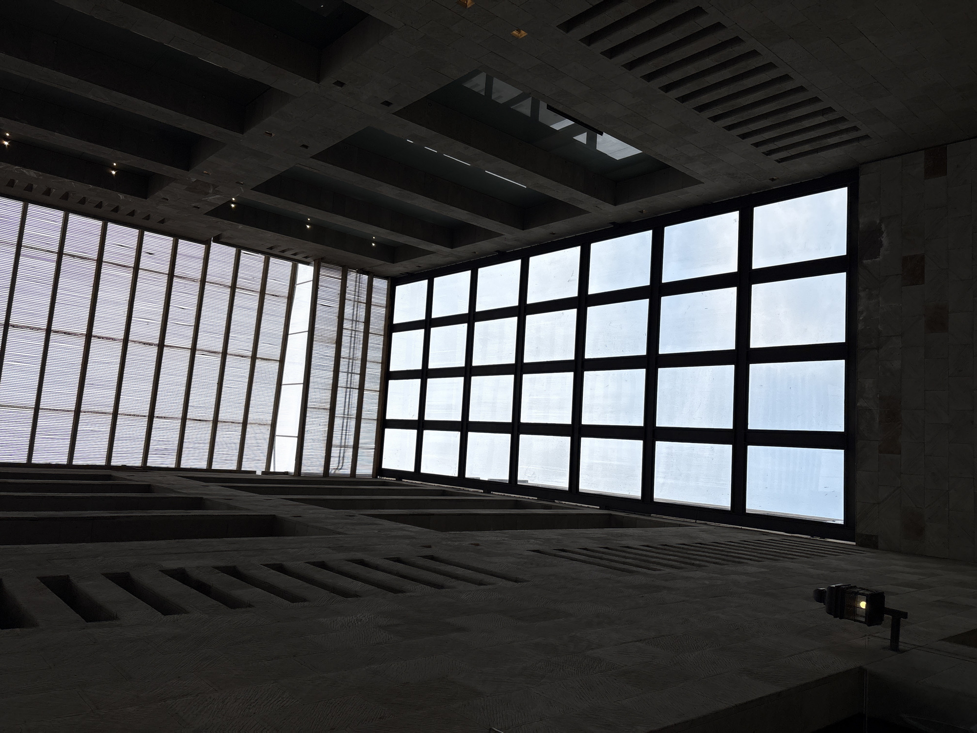 The image shows an interior space with a high ceiling and two large window structures. One window structure is composed of thin vertical slats, while the other is made of square panes. The walls and ceiling are constructed from a textured, light-colored stone. The ceiling has rectangular indentations and a rectangular skylight. On the floor, there are rectangular recesses, and in the lower right corner, a small light fixture is visible. The image provides no information about the location in Ciudad López Mateos, Mexico.