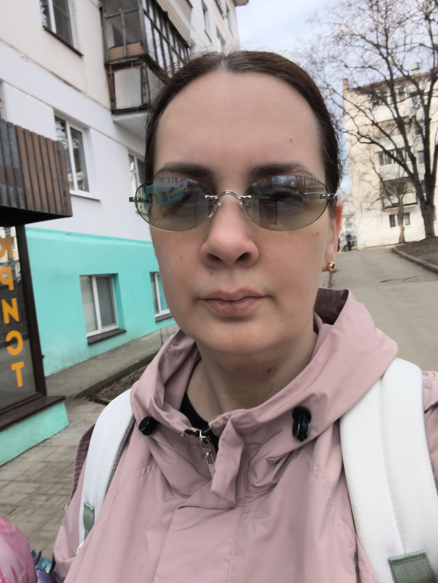 A woman with dark hair pulled back poses for a close-up selfie outdoors in what appears to be an urban or suburban setting. She wears rimless, lightly tinted sunglasses that reflect the surrounding buildings and sky. Her expression is neutral. She is dressed in a light pink hooded jacket worn over a black top, and a white backpack with light green straps is visible on her shoulders. A small gold stud earring is visible on her right earlobe.
The background shows various buildings under an overcast sky. To the woman's left, there is a building with a distinctive bright teal-colored lower facade and white upper sections. Part of a dark brown wooden awning is also visible next to it. On a sign adjacent to this building, partially obscured vertical yellow text, likely Cyrillic, can be seen, with the letters "OT" clearly visible at the bottom. Behind her and to her right, another light-colored building with multiple windows and balconies is present, alongside bare trees with intricate branches, suggesting late winter or early spring. The ground beneath the trees and buildings is a light-colored paved surface, likely a sidewalk or street. The overall lighting is soft, indicating a cloudy daytime.