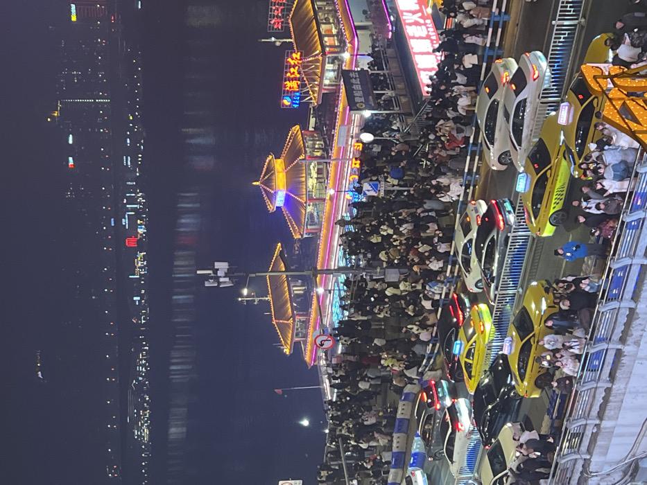 The image captures a densely populated urban waterfront scene at night in Yuzhong District, Chongqing, China. In the foreground, a multi-lane roadway is heavily congested with numerous vehicles, predominantly yellow-green taxis and a variety of sedans, most appearing stationary or moving slowly. Hundreds of pedestrians occupy a promenade directly adjacent to the road, separated by a blue and white striped barrier. In the midground, a large crowd of people gathers around and on platforms associated with several traditional Chinese-style buildings, which are brightly illuminated with prominent yellow, purple, and blue decorative lights. These structures, featuring multi-tiered roofs, are positioned directly alongside a wide, dark body of water. Visible signage on these buildings includes Chinese characters such as "瑞江红" and "渝味老火锅." A "no left turn" traffic sign is affixed to a pole near the illuminated structures. Across the river in the background, the distant cityscape is visible as a collection of illuminated high-rise buildings, their lights reflecting distinctly on the water's surface. Artificial lighting from multiple sources saturates the scene.