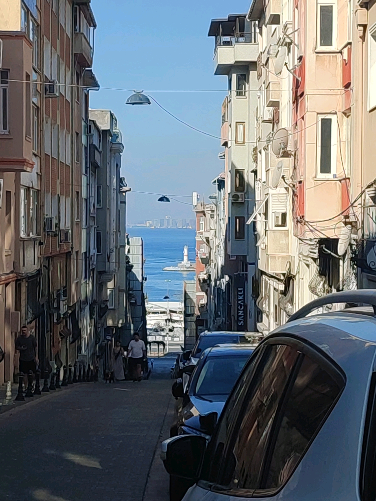 The image depicts a street scene in Kadıköy, Turkey, with buildings lining both sides of a sloping road leading towards the sea. In the distance, a lighthouse is visible on a pier, with a city skyline behind it. The sky is clear and blue.
On the left side of the street, several people are walking. One man, wearing a dark t-shirt and shorts, stands near the side of the road. Further down, a couple is walking with a small dog.
The buildings are multi-story structures with balconies and visible air conditioning units. Satellite dishes are mounted on some of the building facades. Overhead, a wire spans across the street with what appears to be a hanging light fixture.
In the foreground, the hood and windshield of a light-colored vehicle are prominent, reflecting the buildings across the street. A dark-colored car is parked behind it. The road surface is paved. The overall impression is of a sunlit day in an urban environment with a view of the water.