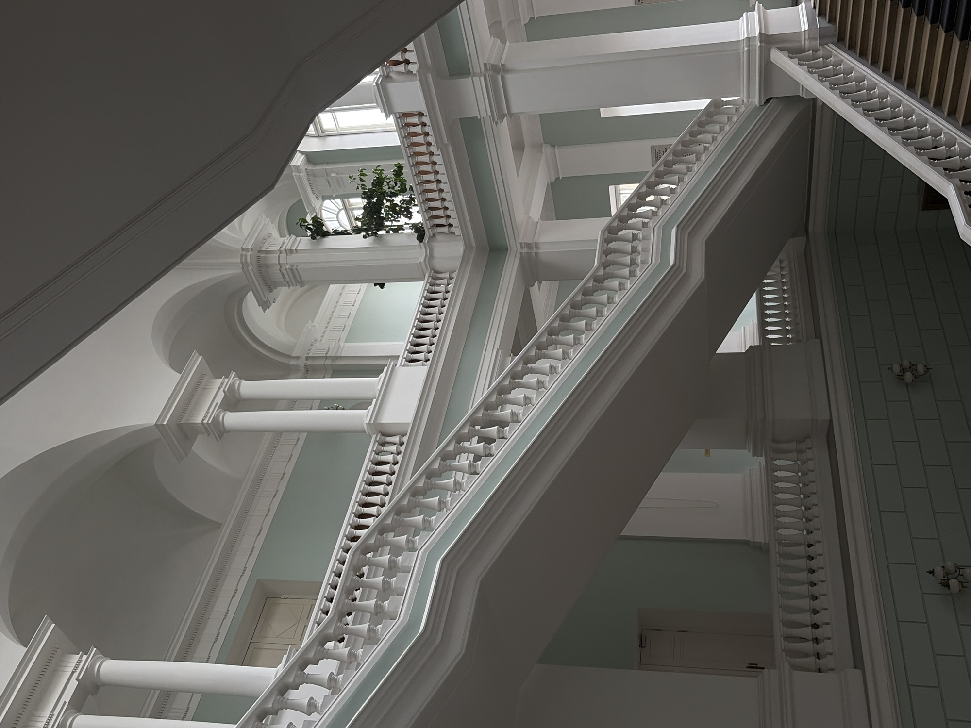 The image depicts a grand interior staircase with white balustrades and light blue walls. The staircase is a prominent feature, ascending through multiple levels of the building. Arches and columns contribute to the classical architectural style. A small tree with green foliage is visible on an upper landing. The scene appears to be a public building, possibly a museum or historical institution, given the opulence and scale. The lighting is soft and diffused, suggesting an interior space illuminated by natural light from windows, some of which are visible. No people are present in the image. The overall impression is one of elegance and architectural grandeur.