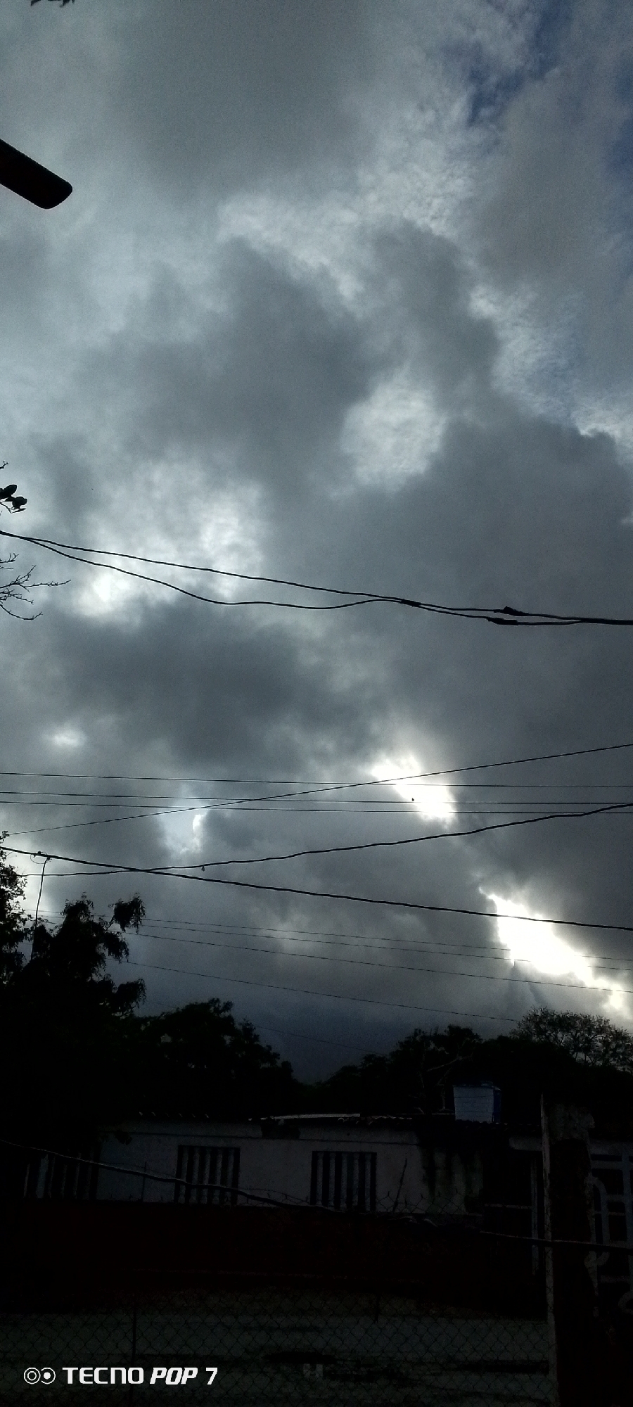 A vertical, low-angle shot captures a dramatic sky filled with dark, heavy clouds. Faint breaks in the cloud cover allow shafts of bright light to penetrate, creating a somewhat ominous atmosphere. Several power lines crisscross the frame horizontally, silhouetted against the sky. In the lower portion of the image, the tops of dark, silhouetted trees are visible, with a partially visible building featuring barred windows. The bottom edge of the frame shows a dark fence and the text "TECNO POP 7." The overall scene suggests a potential storm or overcast weather conditions in a semi-urban or rural environment, likely Altagracia, Venezuela, given the watermark. There are no people or distinct actions depicted.