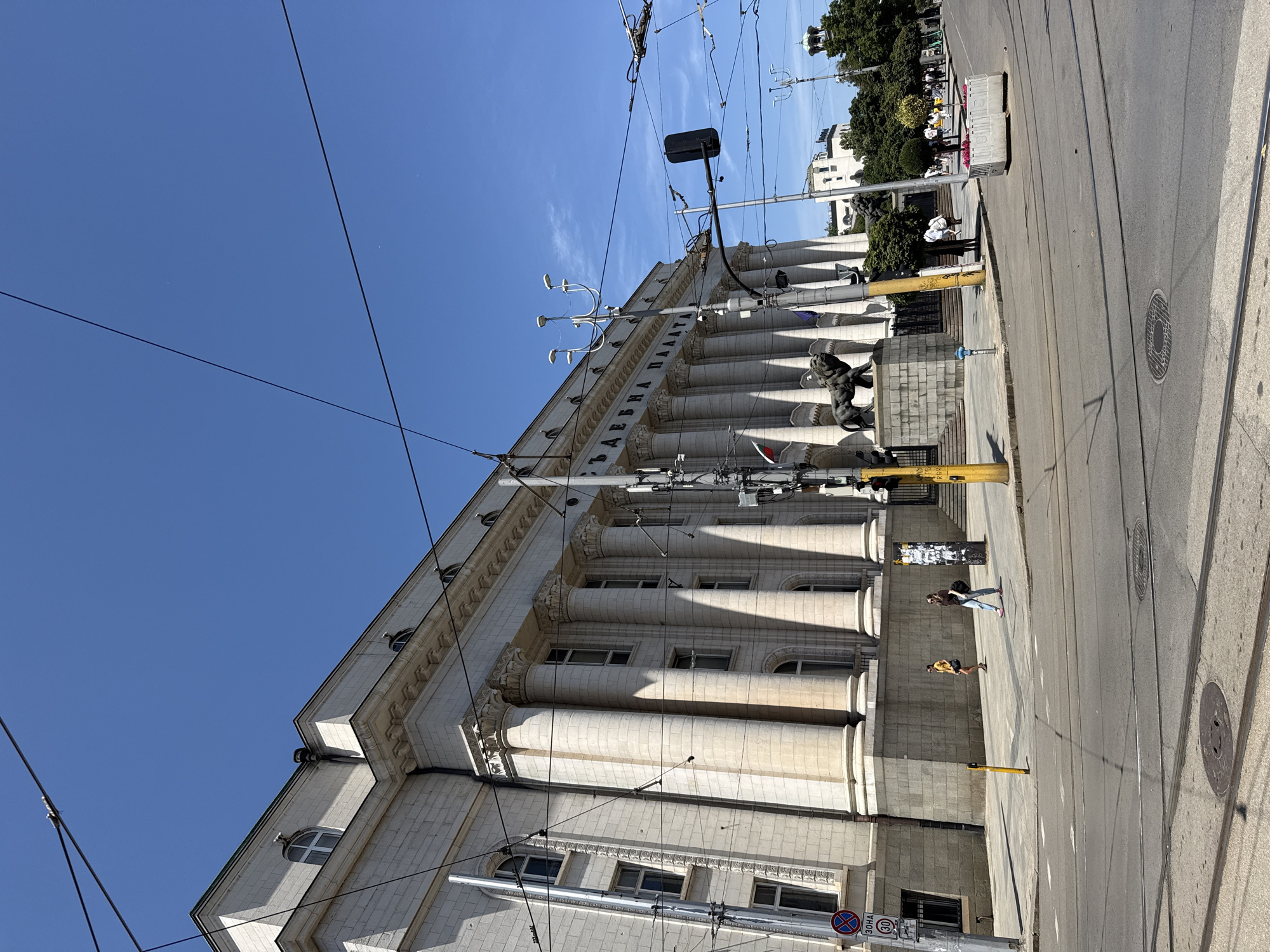 The image depicts a street scene in Sofia, Bulgaria, dominated by a large, neoclassical building with prominent columns. The building appears to be the Palace of Justice, judging by the lion statues at its entrance. A clear blue sky with some wispy clouds serves as the backdrop. Overhead, numerous overhead wires, likely for trams or trolleybuses, crisscross the frame.
In the foreground, a paved street with tram tracks is visible. The pavement on the sidewalk is also visible. Several people are present on the sidewalk. Two women are walking, one wearing a yellow top and dark shorts, the other in a dark top and jeans. Further down the street, a group of people are gathered.
Key architectural features of the building include a long colonnade, arched windows, and decorative moldings. A traffic light is mounted on a pole, and a circular sign with a "30" inside is affixed to another pole. Yellow metal poles are strategically placed along the sidewalk. In the distance, other buildings and trees can be seen. The overall impression is a sunny day in a city with a rich architectural heritage and active public transportation system.