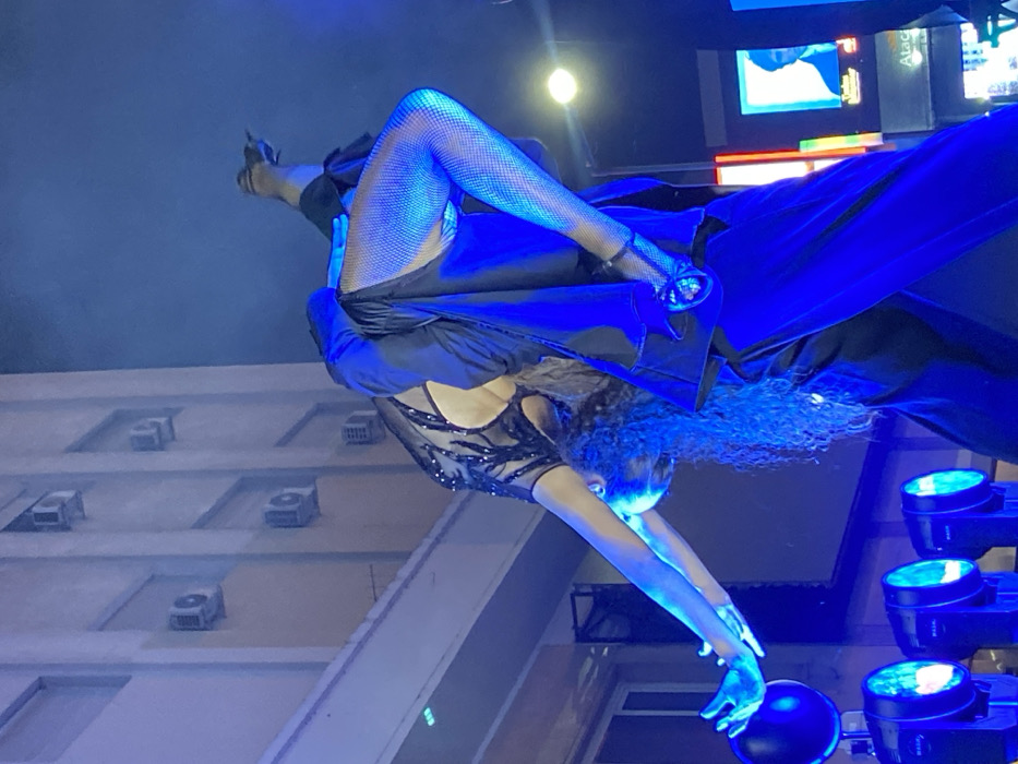 A contortionist performs in front of a building and stage lighting. The performer, wearing fishnet stockings and a sequined bodysuit, bends backward with arms outstretched toward black spheres. A second individual, partially visible, is wearing a black suit. The surrounding environment includes an exterior building facade with multiple air conditioning units. The scene is bathed in blue stage lighting.