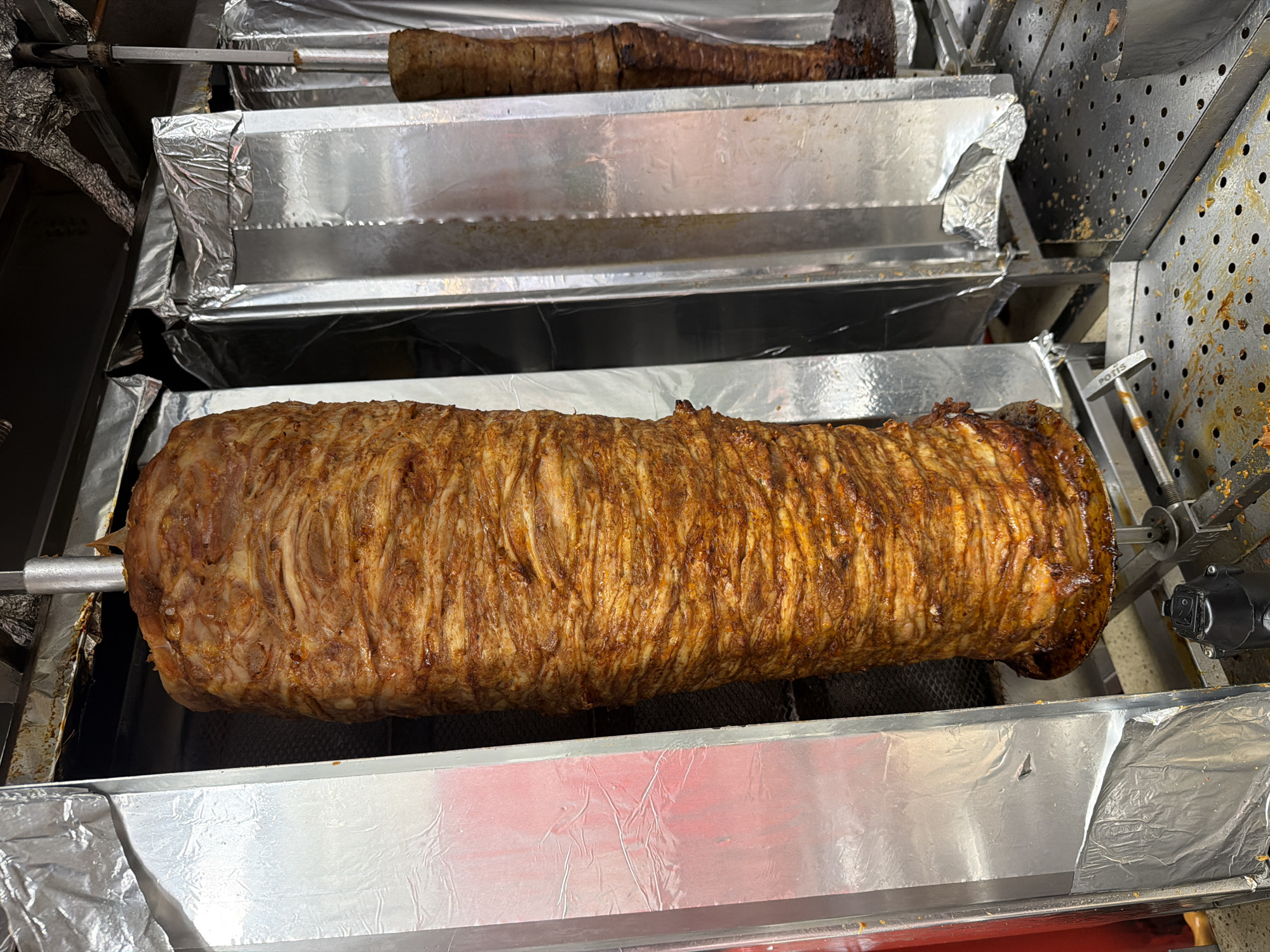 The image shows a vertical spit of meat cooking in a döner kebab machine. The meat is thinly sliced and stacked, with a visible browned exterior indicating it is being roasted. The machine is positioned in what appears to be an outdoor food stall or restaurant kitchen. The background reveals more of the cooking apparatus, including another, smaller spit of meat and reflective surfaces, likely aluminum foil, used for heat reflection and insulation. The overall scene suggests a food preparation environment, possibly a street food vendor. The image is taken at ground level and is well-lit, focusing on the meat.
