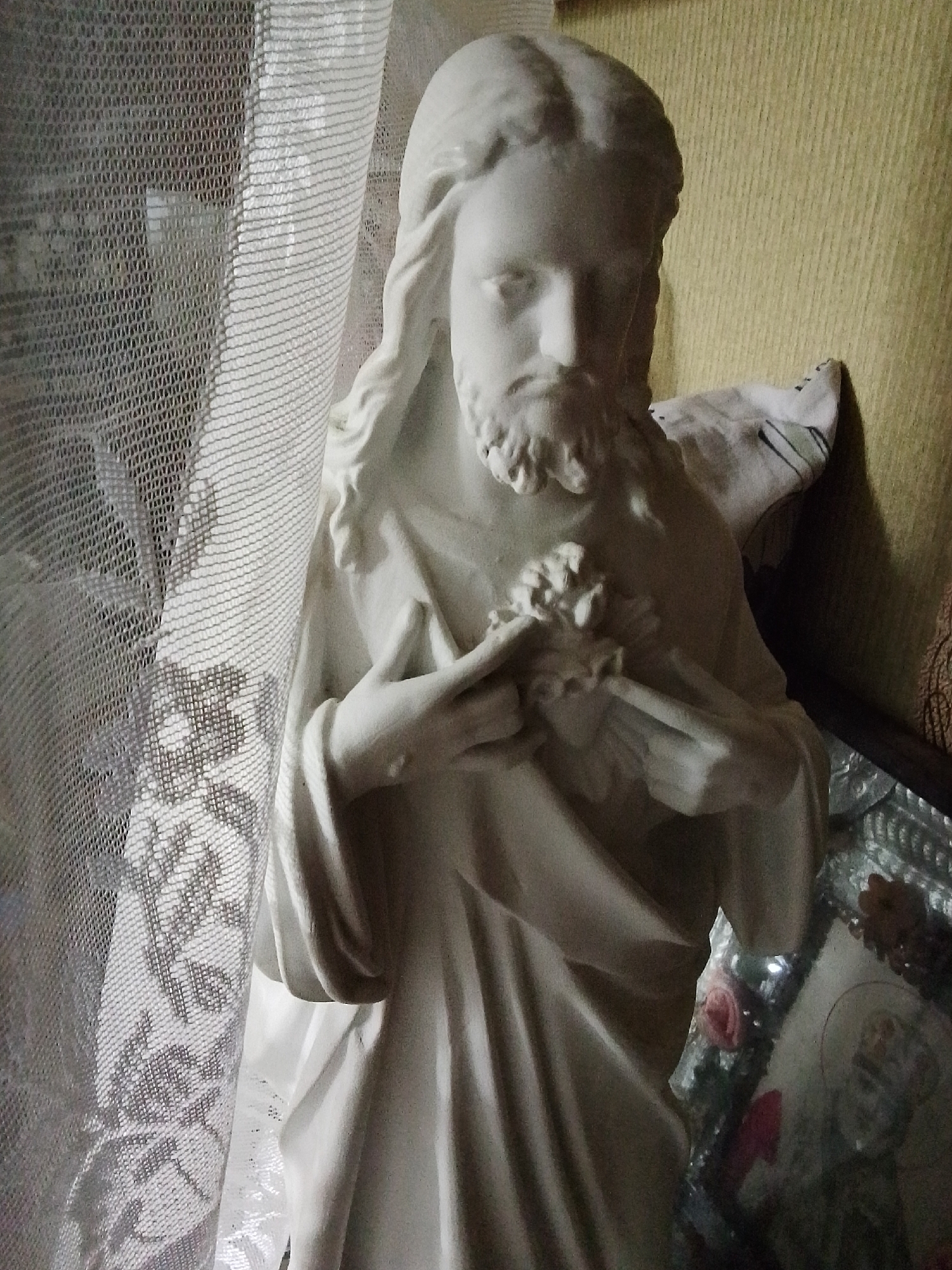 The image presents a white statue of Jesus Christ, captured from a slightly elevated, eye-level perspective. The statue depicts Jesus with long hair and a beard, and his hands are clasped over a Sacred Heart emblem on his chest. The material appears to be plaster or a similar smooth, light-colored substance.
To the left of the statue, a translucent white lace curtain or fabric hangs, revealing a floral pattern through its mesh. This curtain partially obscures the background on that side. Behind the upper right portion of the statue, a wall with a distinct yellowish-brown color and a vertical textured, possibly ribbed, surface is visible. A folded piece of white and striped fabric rests against this wall, near the statue's right shoulder.
Below the statue and to the right, a reflective surface, likely a decorative tray or a framed mirror, displays several small items. These include a circular religious icon or picture and artificial flowers, notably a pink rose and a red bloom.
The scene suggests an indoor domestic setting, possibly a corner dedicated to religious devotion within a home in Khlevnoye, Russia. The lighting is soft and ambient, contributing to a calm and contemplative atmosphere. No specific activity is taking place, as the image captures a static arrangement of religious artifacts. There is no visible text within the frame.