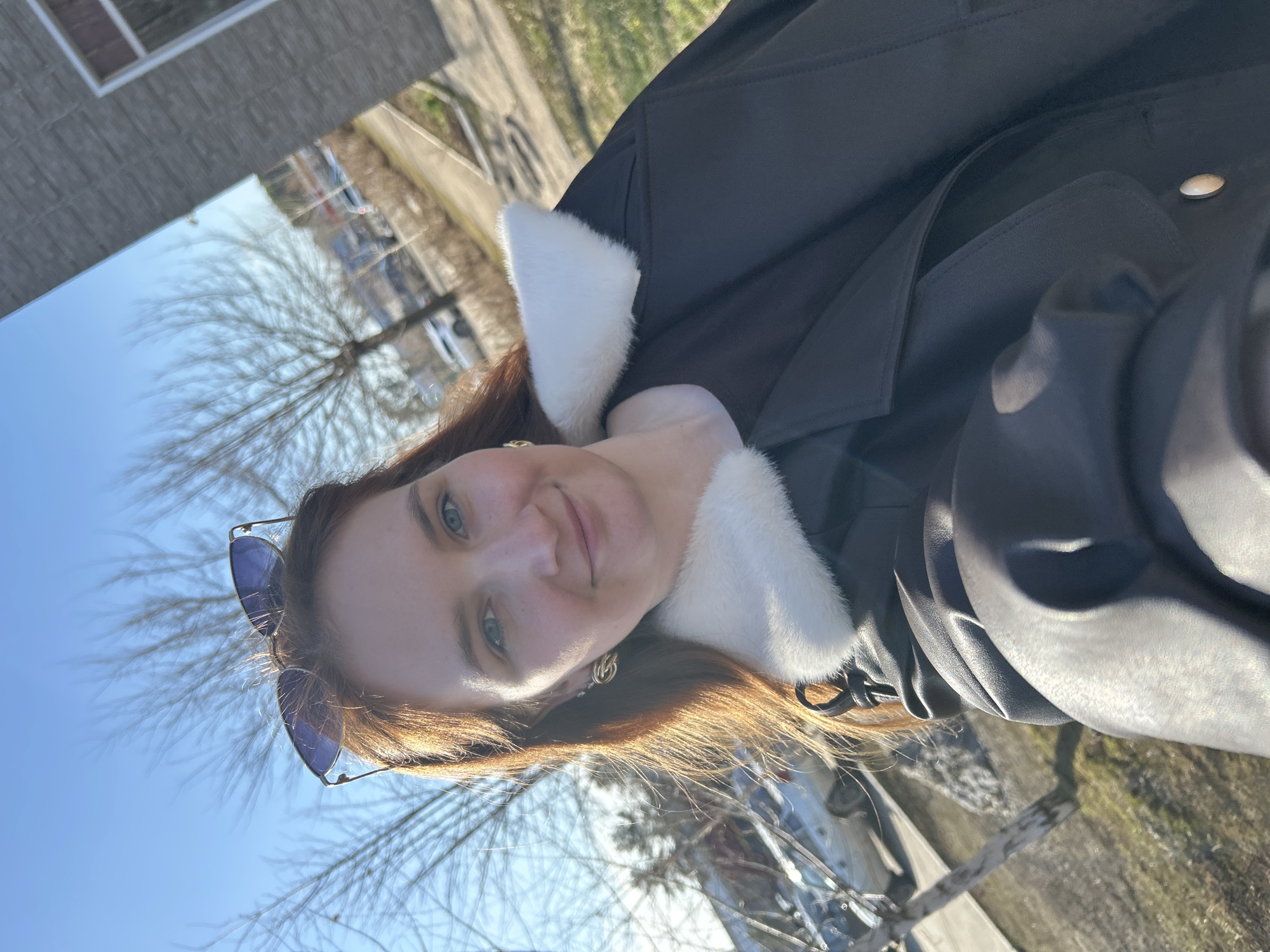 A woman with red hair and blue eyes is posing for a selfie outdoors. She is wearing sunglasses perched on her head and a black coat with a white fur collar. Her hair is blowing in the wind, and she is smiling slightly. The background shows a city street with trees, buildings, and a clear blue sky. The location appears to be Chaikovsky, Russia.