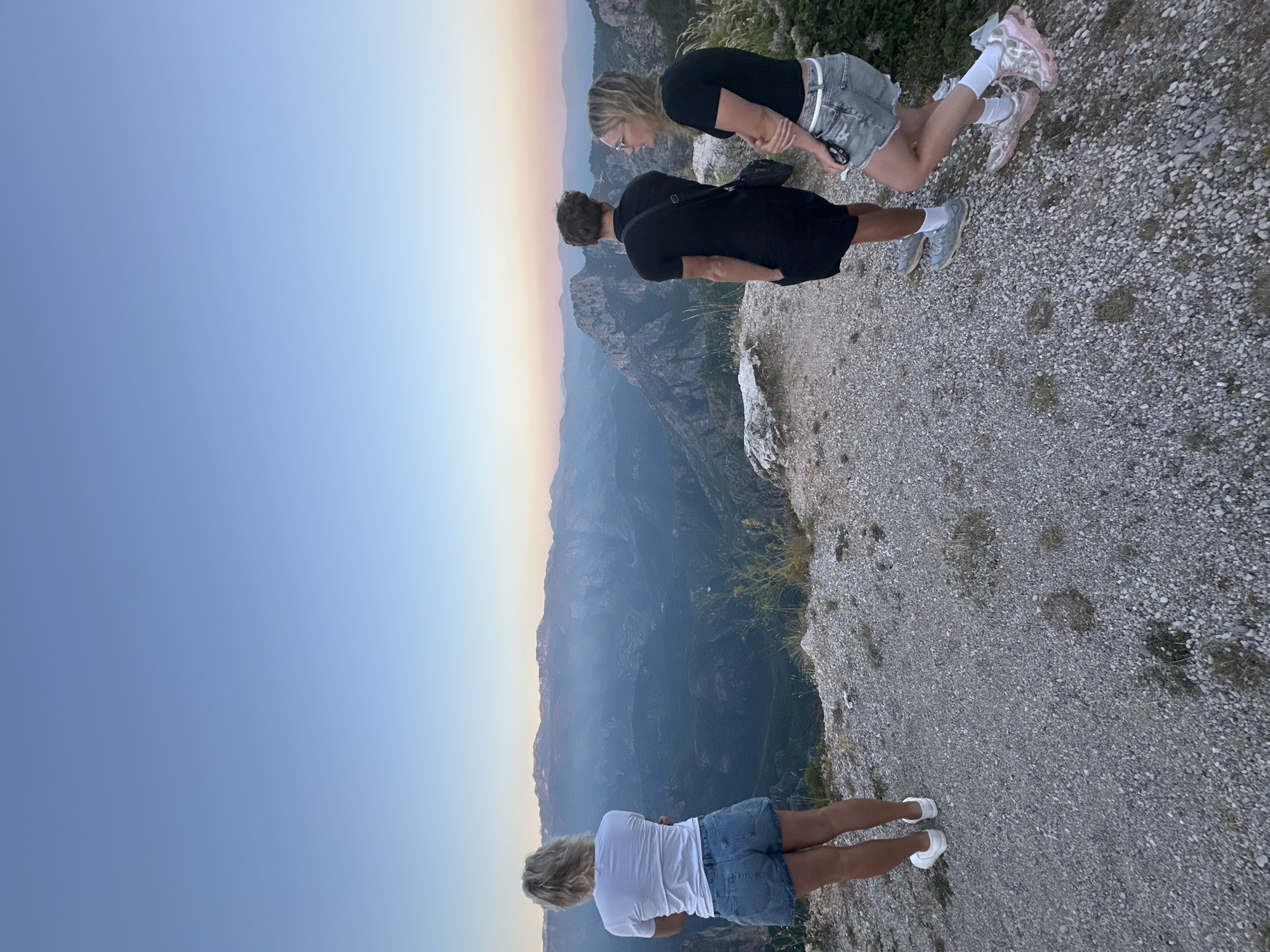 The image depicts three individuals standing on a rocky, elevated viewpoint overlooking a vast mountain range at dusk. The sky is a gradient of soft blues and oranges, indicating the setting sun.
In the foreground, a woman with blonde hair stands with her back to the camera, wearing a white t-shirt and denim shorts. To her right, two other individuals, a man and a woman, are also facing the vista. The man is wearing a black t-shirt and shorts, with a bag slung across his chest. The woman is wearing a black t-shirt and denim shorts. They are all positioned near the edge of the cliff, engaged in observing the landscape.
The environment is characterized by rugged, mountainous terrain with sparse vegetation. The rocky ground in the foreground contrasts with the deep valleys and layered mountains stretching into the distance, partially obscured by a hazy atmospheric effect. The overall impression is one of a serene, natural landscape viewed from a high vantage point during the twilight hours.
While the image captures a scenic view, the provided information about the location being Aranđelovo, Bosnia and Herzegovina, cannot be verified solely from the visual content of the photograph.
