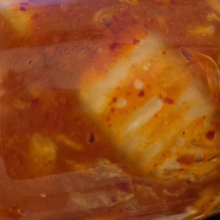 The image presents a magnified view of a food substance enclosed within a transparent container. The substance is characterized by a dominant reddish-orange liquid or paste, which contains numerous irregular, pale-yellowish-white solid components. These solid components exhibit a fibrous or leafy texture. Dispersed throughout the liquid and adhering to the solids are numerous small, dark-red particulate specks, consistent with spices or chili flakes. The container material appears transparent and smooth, reflecting ambient light. No discernible human figures, actions, interactions, or external environmental details are present within the captured frame. The location in the city of Strasbourg, France, cannot be determined from the visual information provided.
